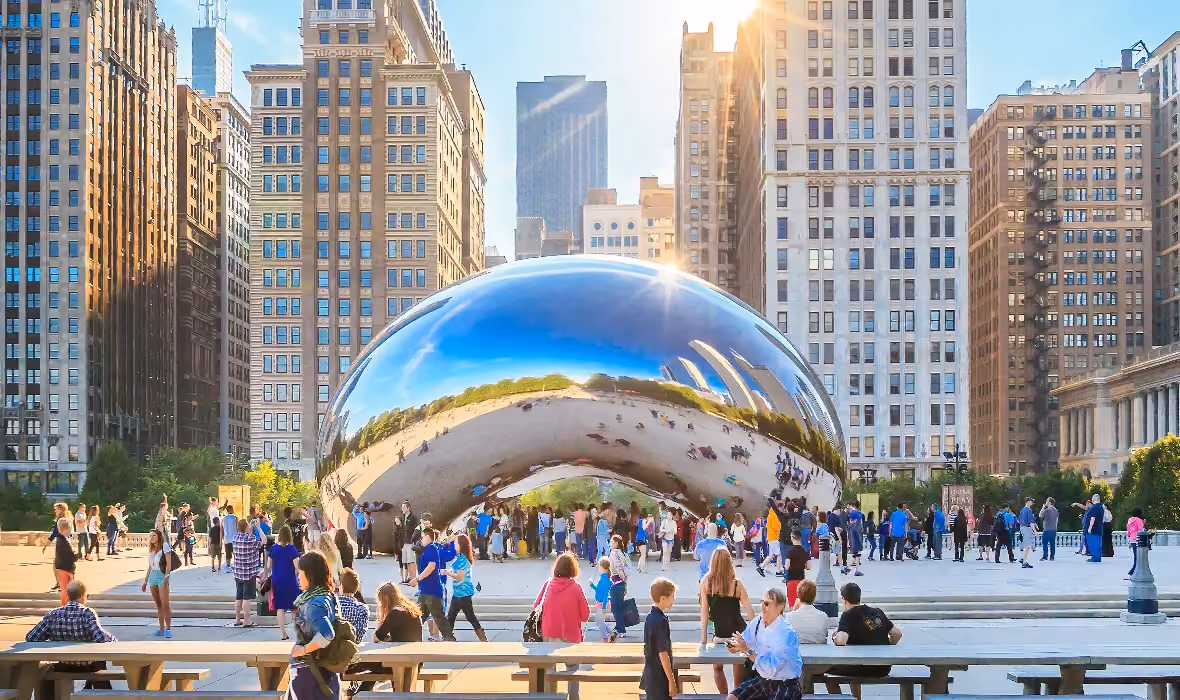 Crowds of people gathered around Cloud Gate, a large reflective bean-shaped sculpture, in a city plaza surrounded by tall buildings on a sunny day.