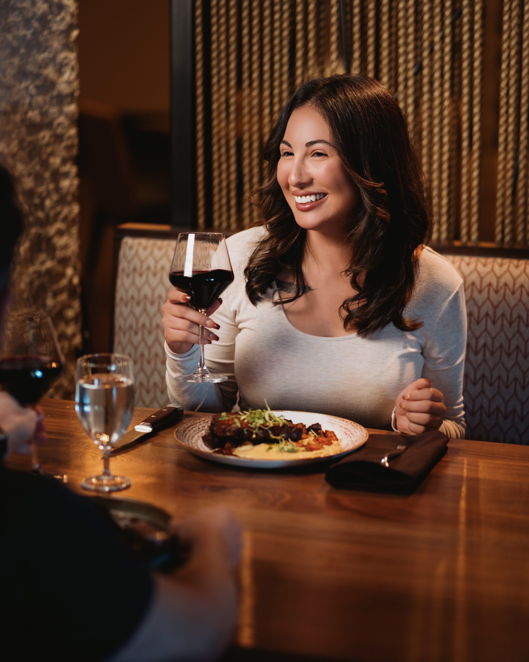 Smiling woman holding a glass of red wine at a restaurant table with a meal in front of her.