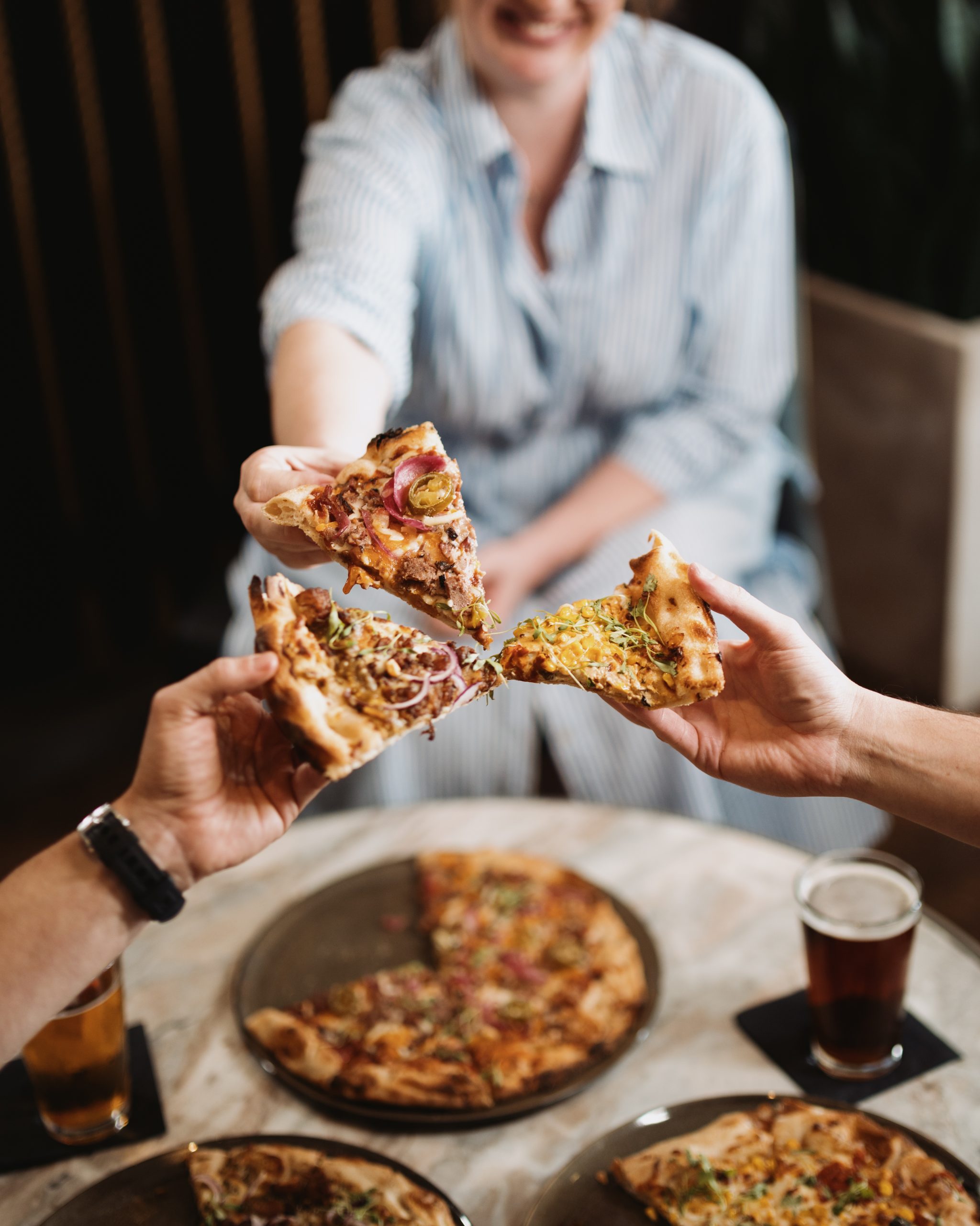 Three people clinking slices of different pizzas over a table with drinks and pizzas.