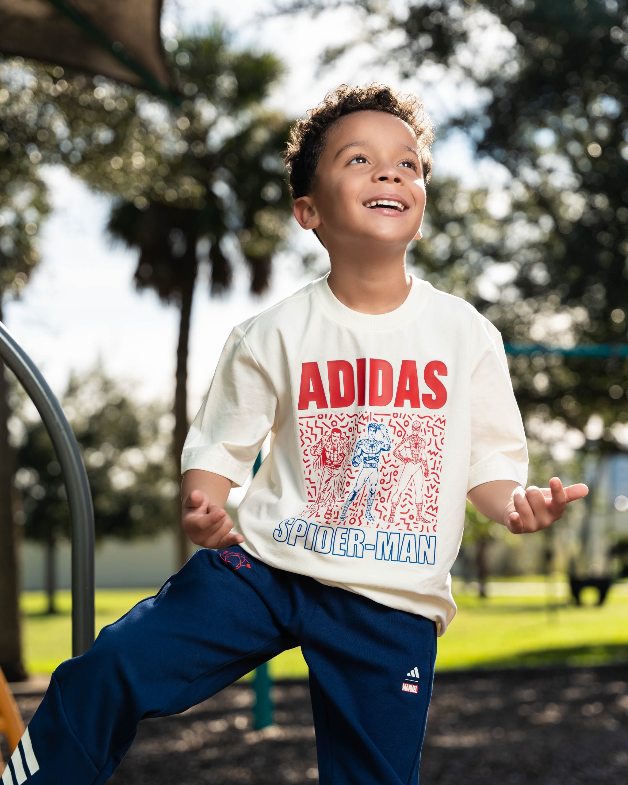 Smiling boy outdoors wearing white Adidas Spider-Man t-shirt and navy Adidas Marvel pants.