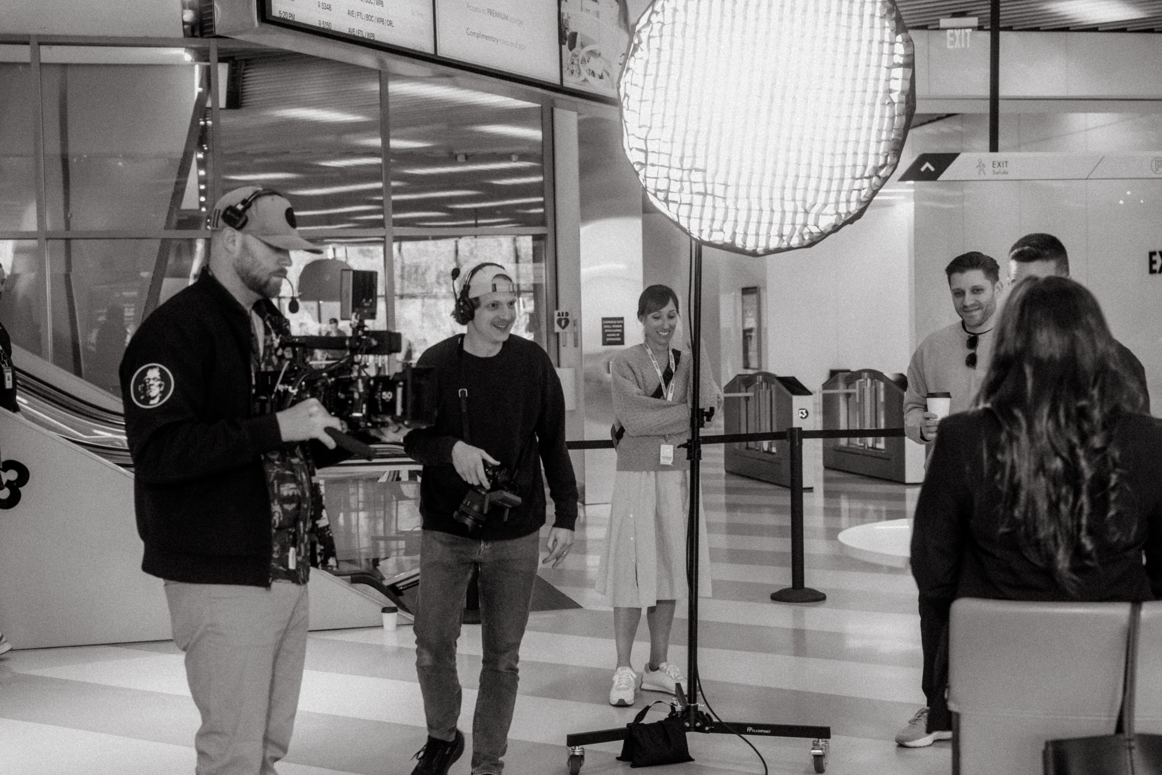 Film crew with cameras and lighting equipment filming two people sitting while others watch.