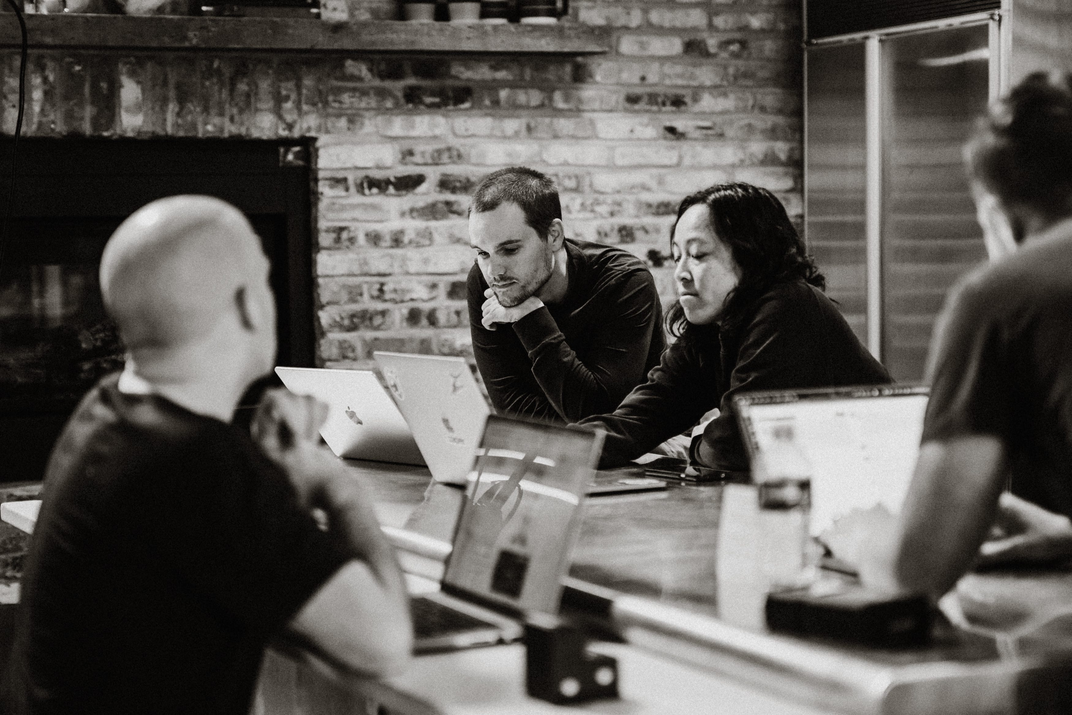 Four people sitting around a table with laptops, two focused on a screen together, in a brick-walled room.