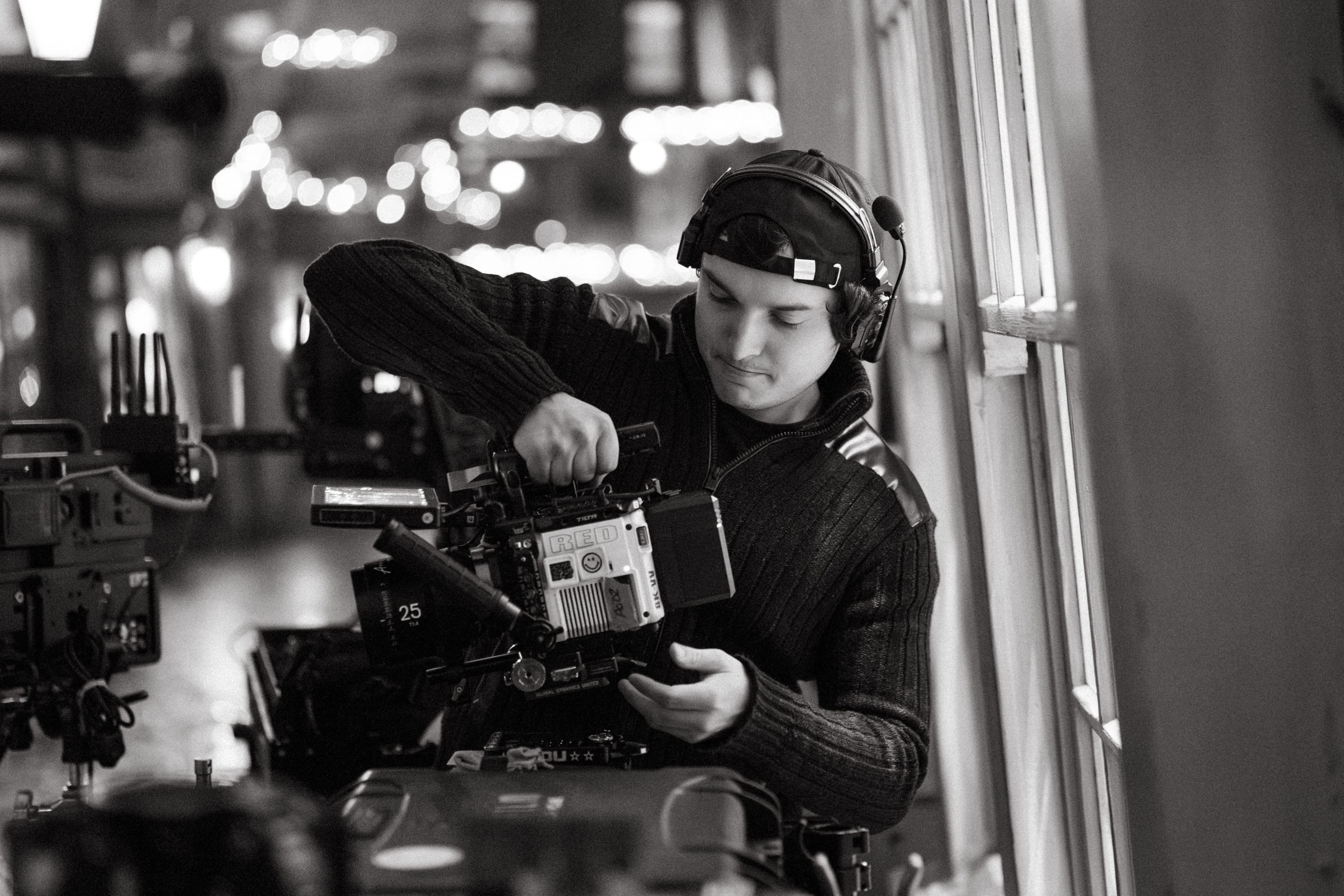 Man wearing headset adjusts a professional video camera indoors near a window.