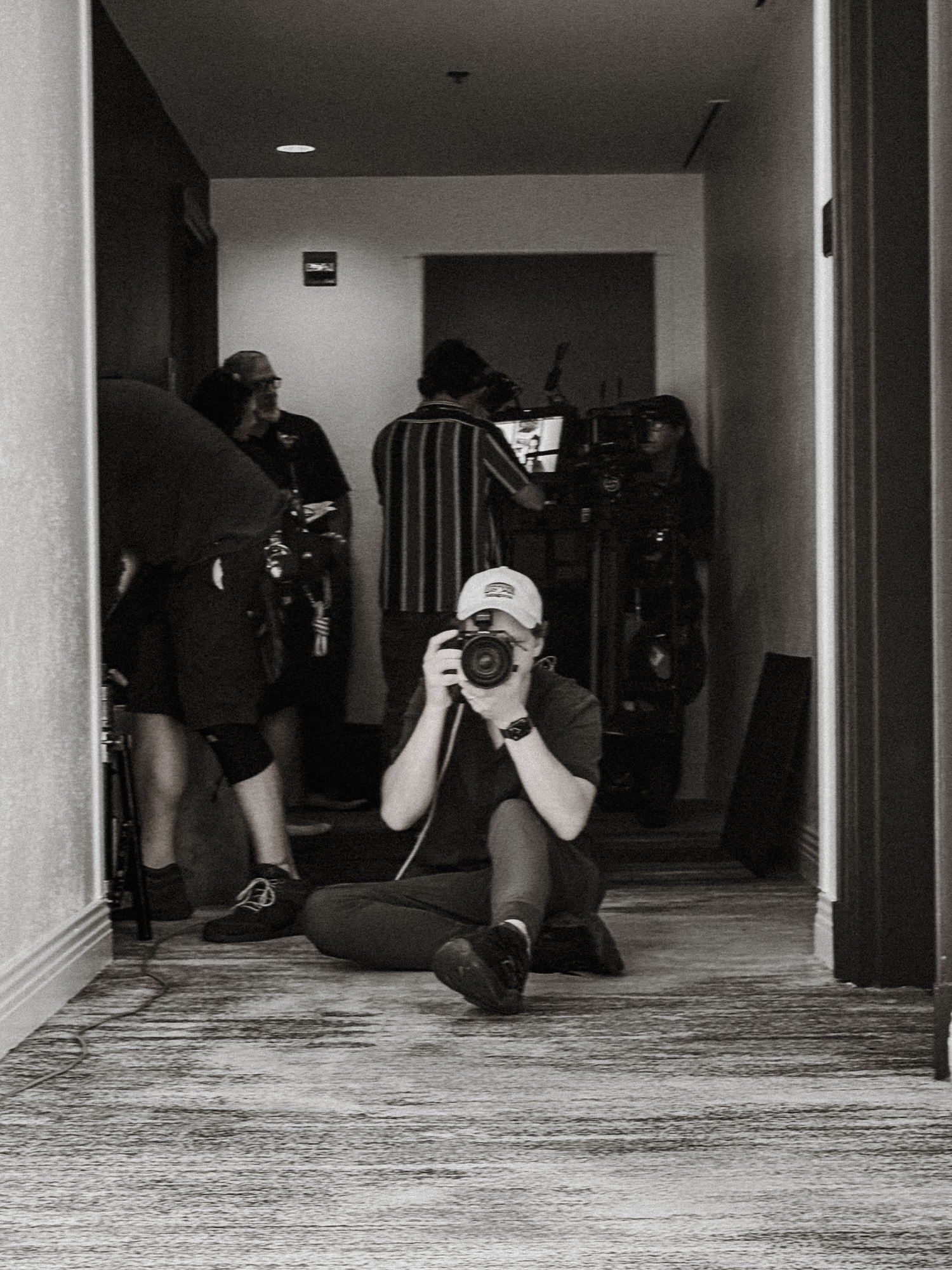 Person seated on hallway floor taking a photo with camera, others standing and filming in background.