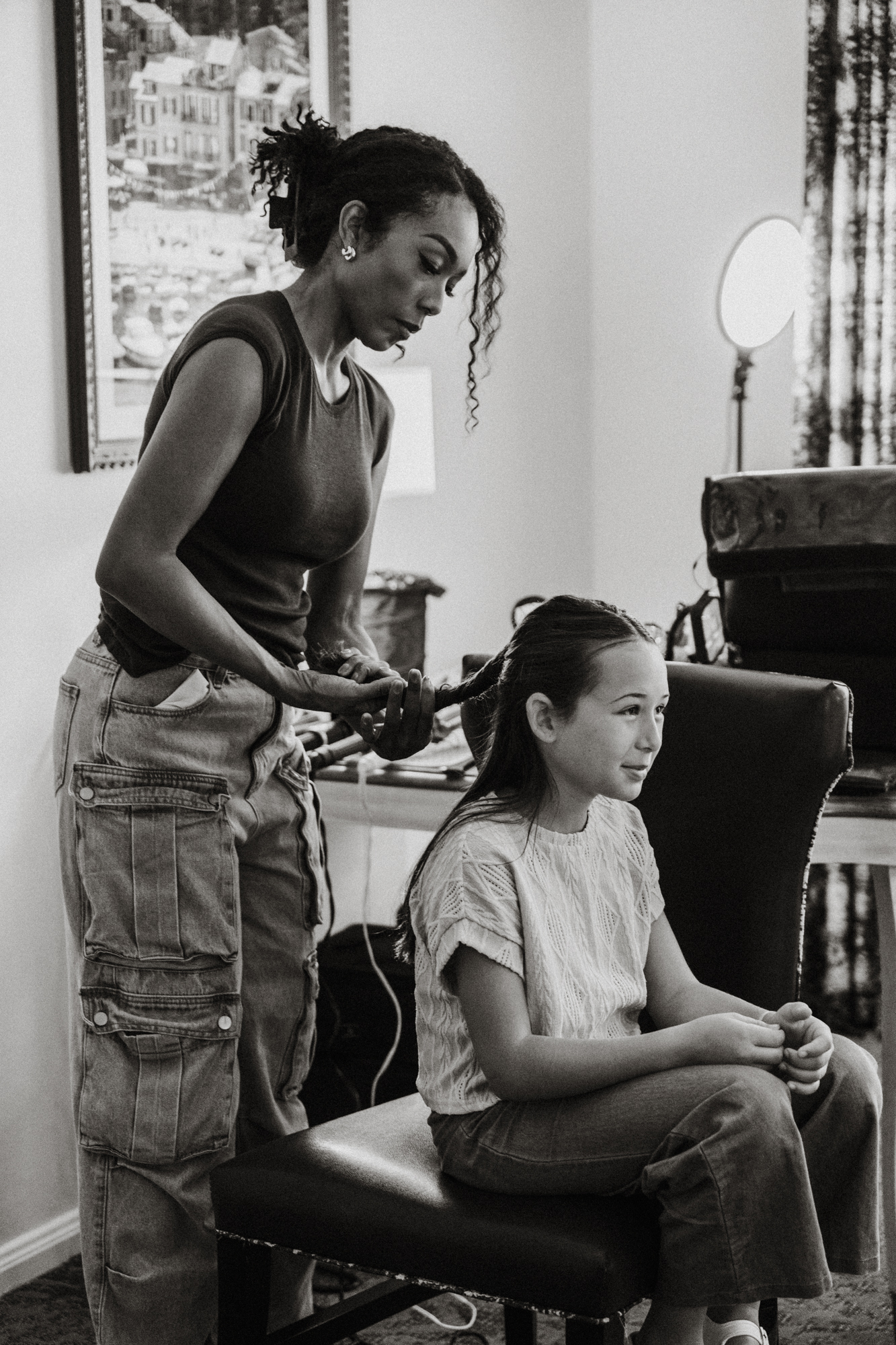 Woman styling a young girl's hair as the girl sits on a chair indoors.