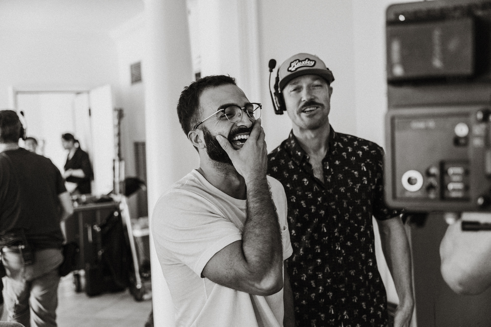 Two men laughing on a film set, one with glasses and a beard, another wearing a headset and cap.