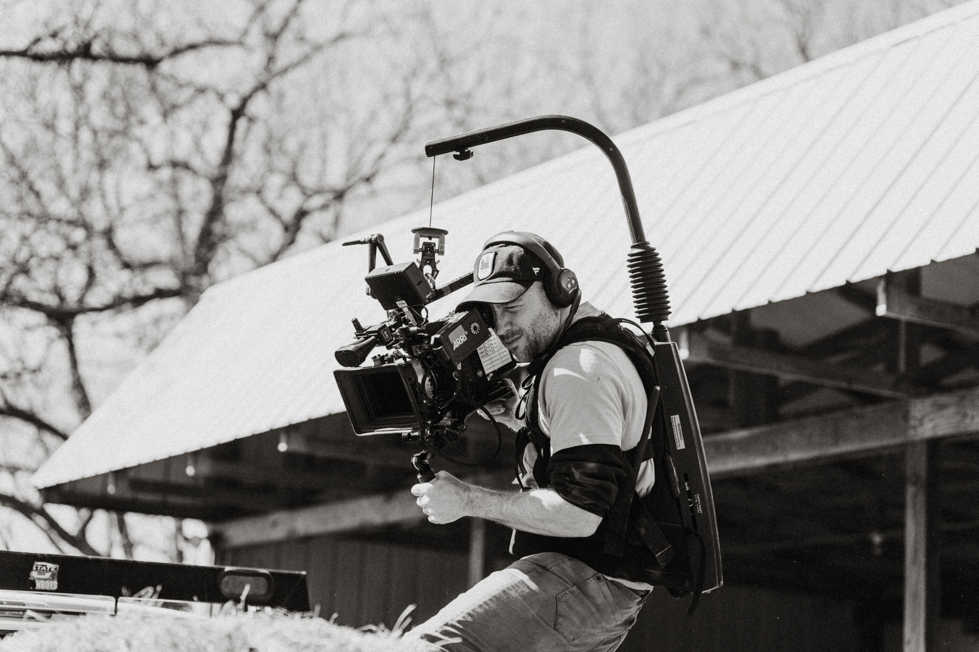 Filmmaker wearing stabilizer rig and headphones operating a professional ARRI camera outdoors near barn.