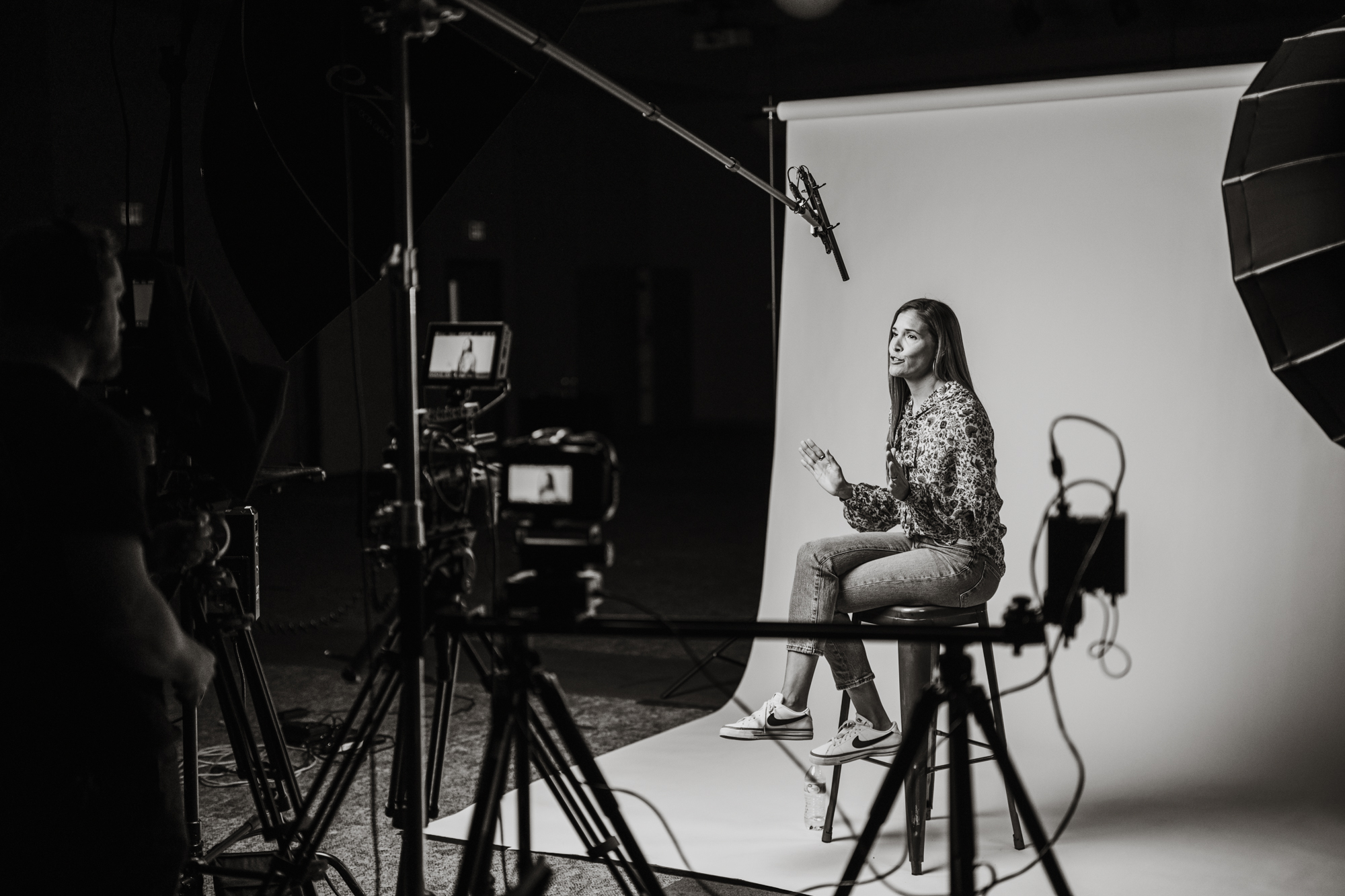 Woman sitting on a stool in a studio, speaking in front of cameras and lighting equipment.