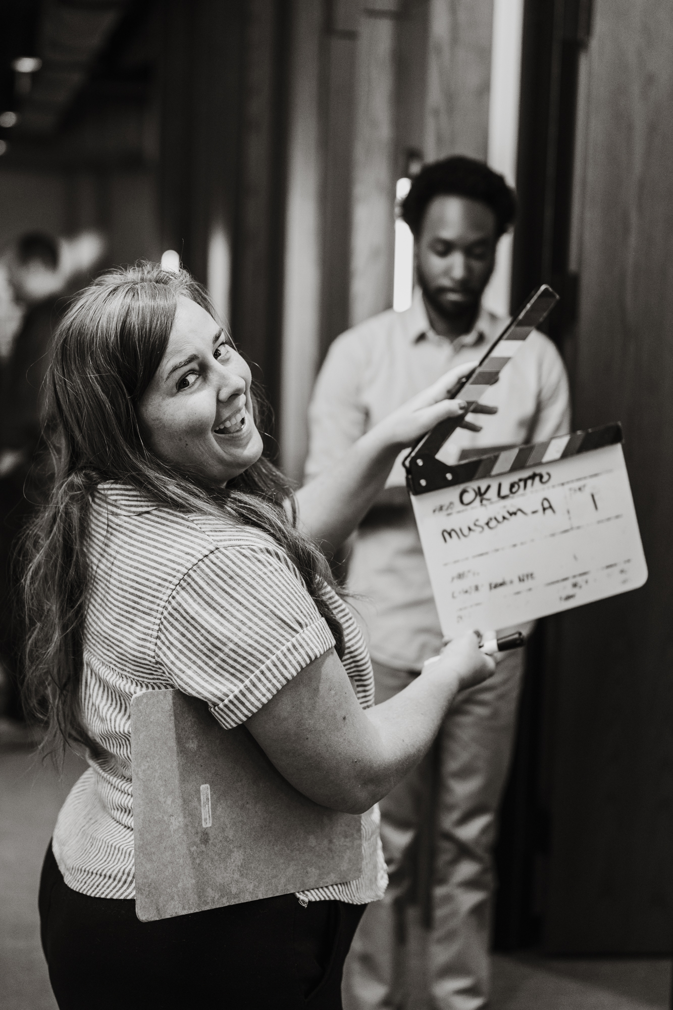 Smiling woman holding a film clapperboard and clipboard with a man blurred in the background.