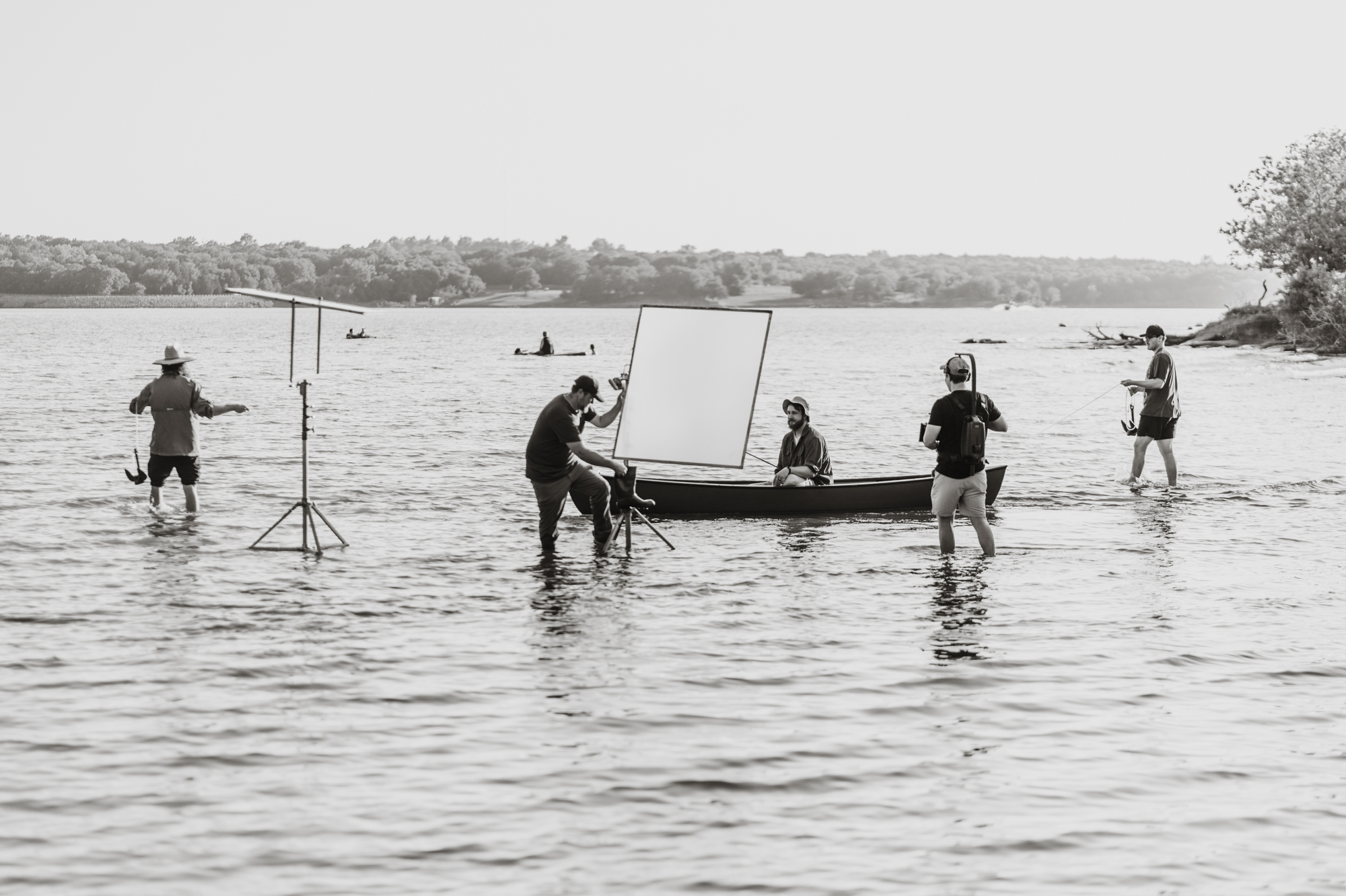 Film crew working in shallow lake water around a canoe with equipment and a seated actor.