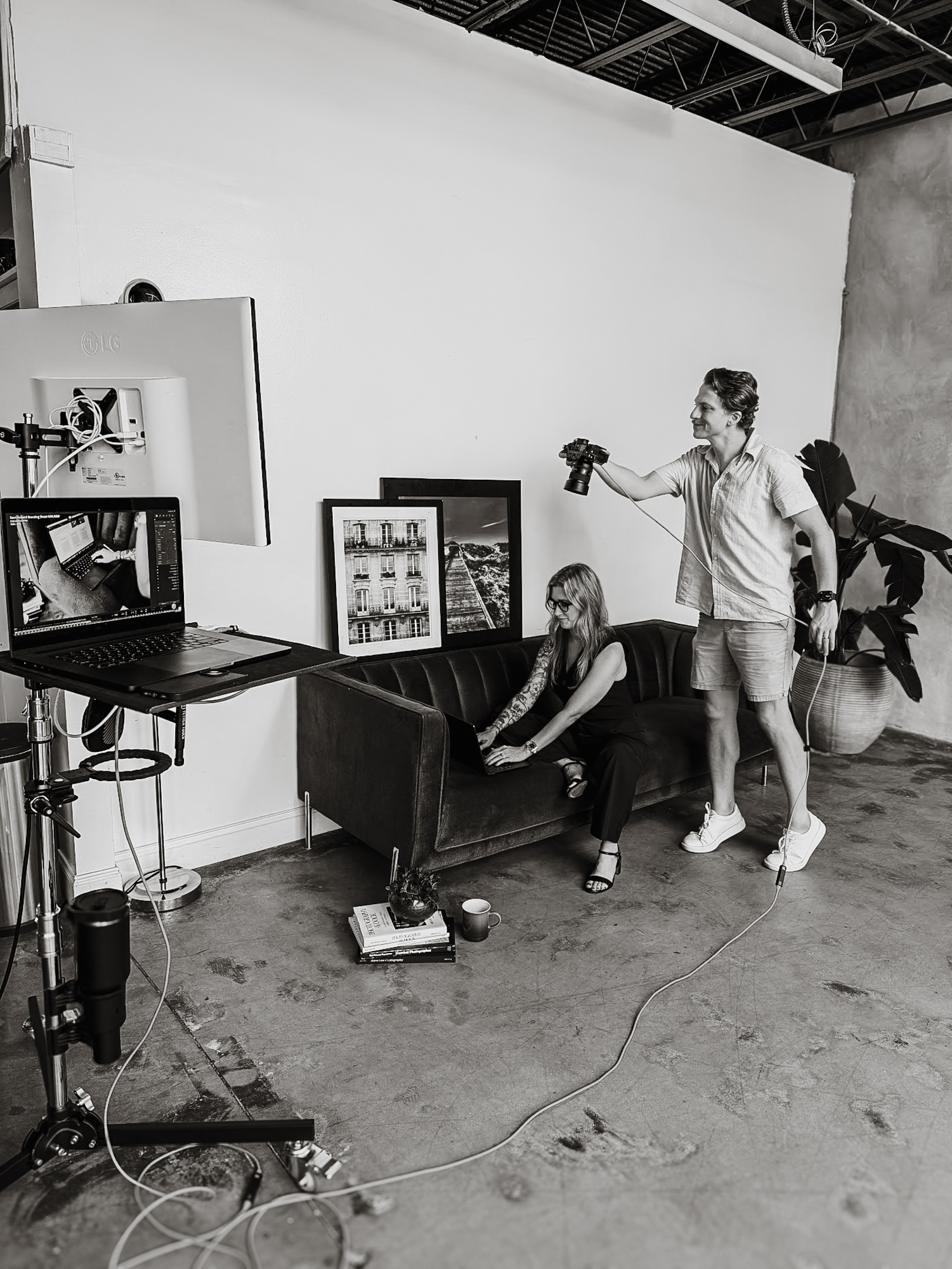 Man holding camera and woman sitting on couch working on laptop in a photo studio setting.