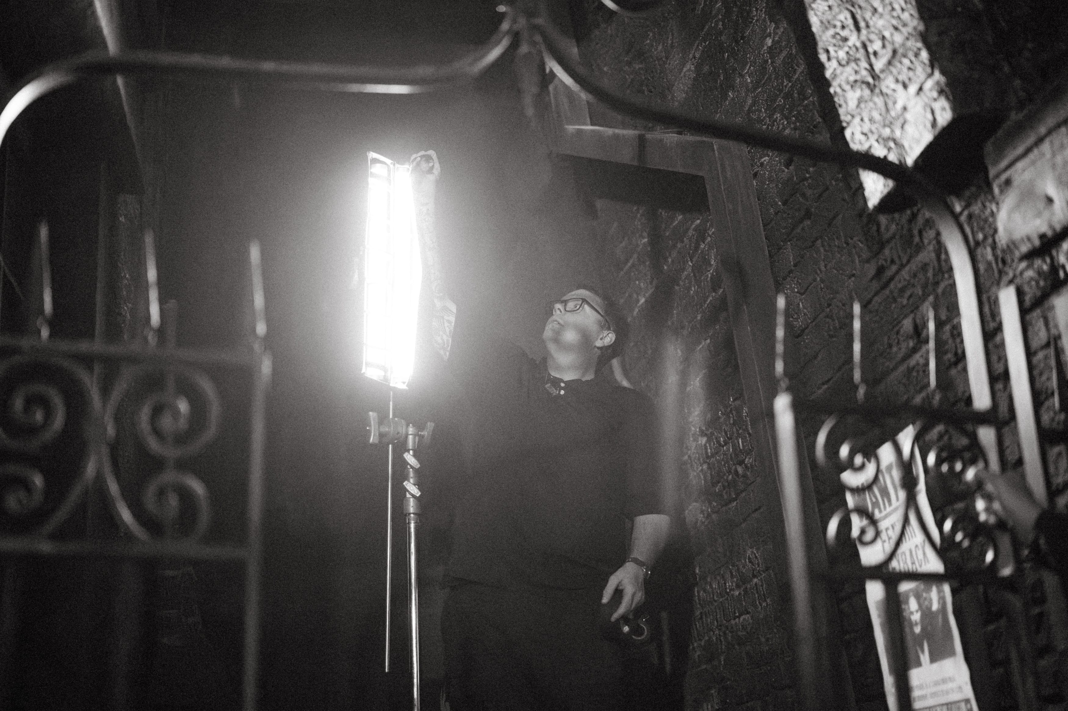 Man adjusting a bright light panel on a stand beside a brick wall in a dim setting.