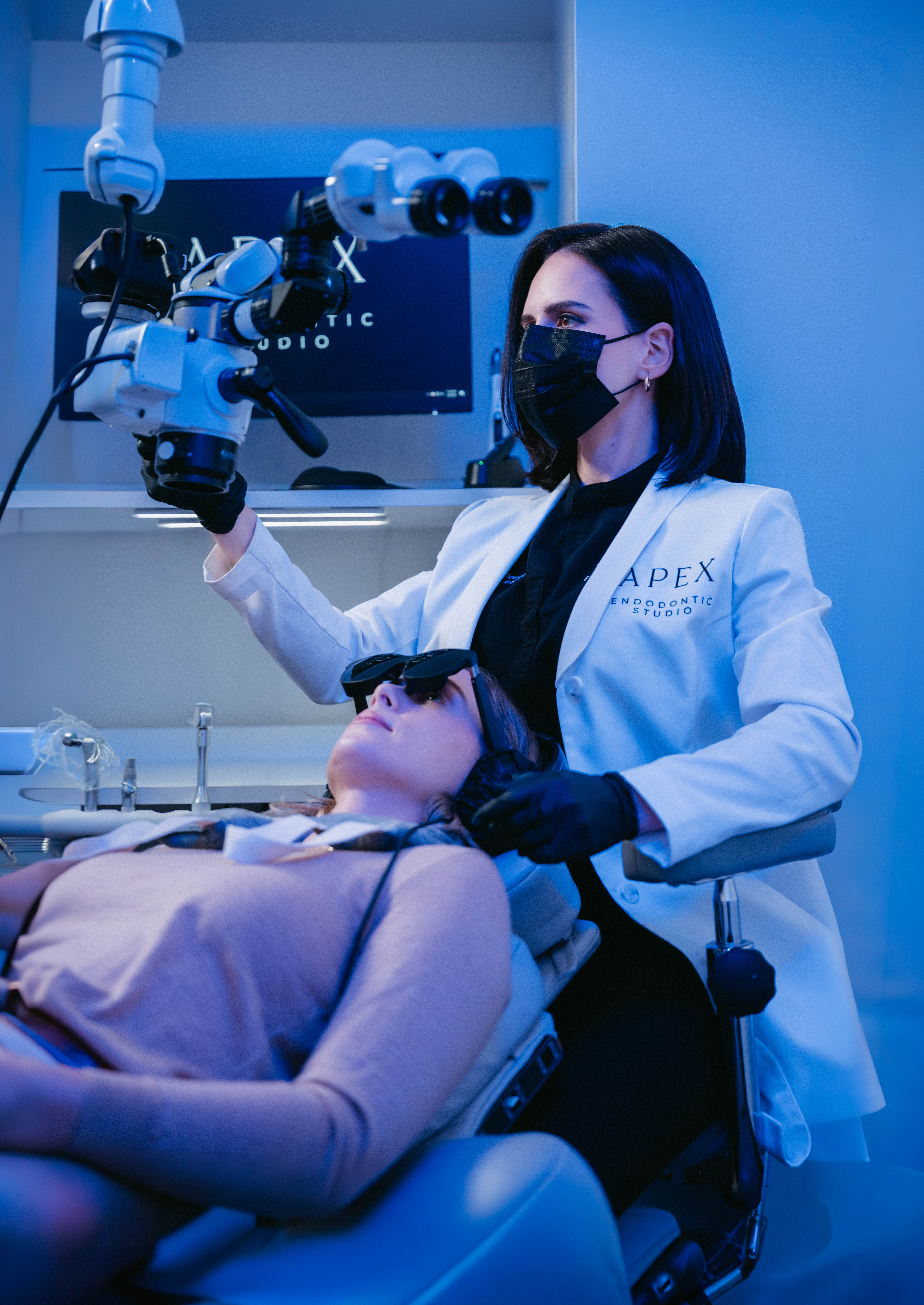 Dentist wearing white coat and mask uses microscope equipment while patient reclines in chair with protective glasses.