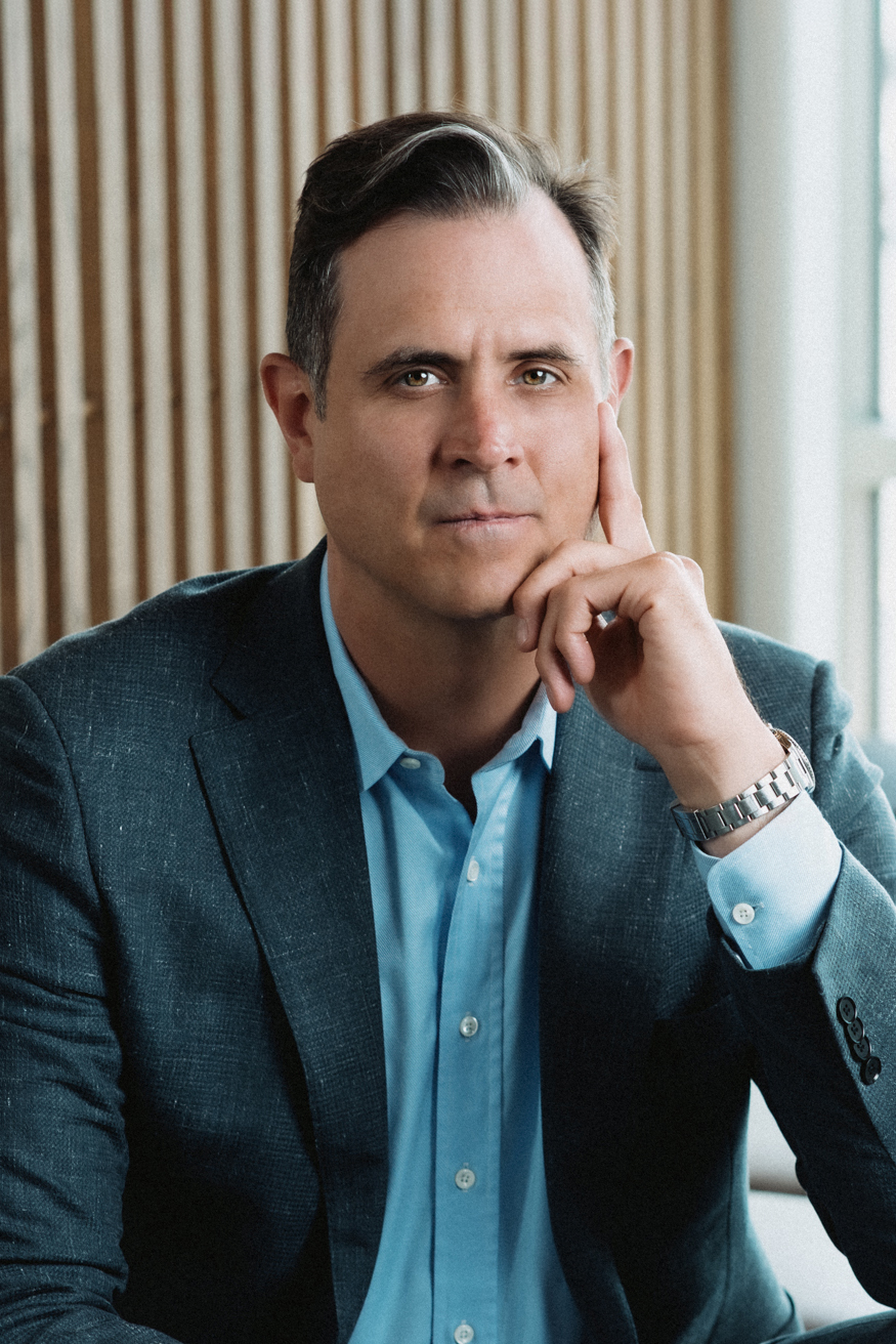 Man in blue shirt and dark blazer resting his face on his hand looking thoughtfully at the camera.