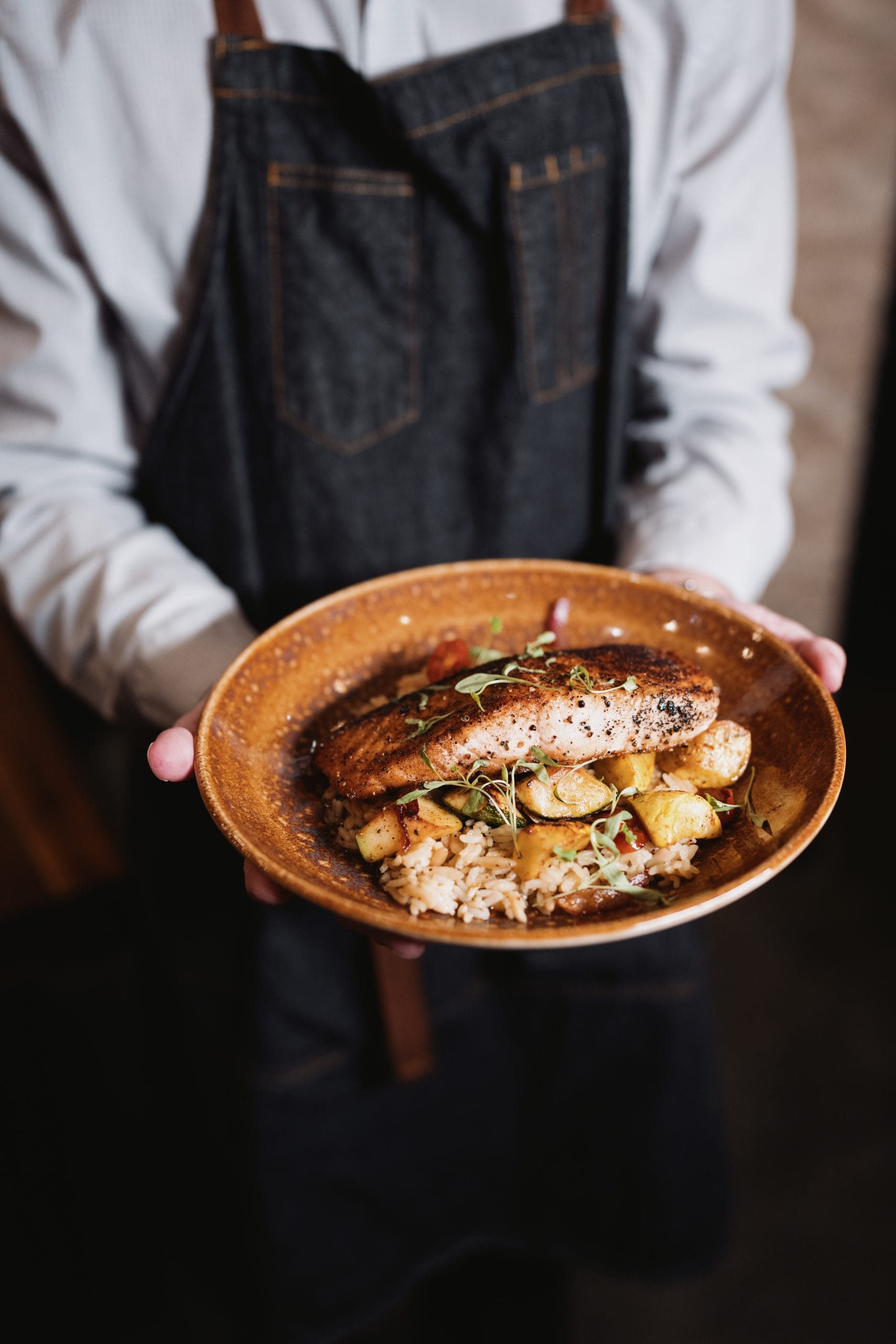 Person in apron holding a plate with grilled salmon, rice, roasted veggies, and microgreens.