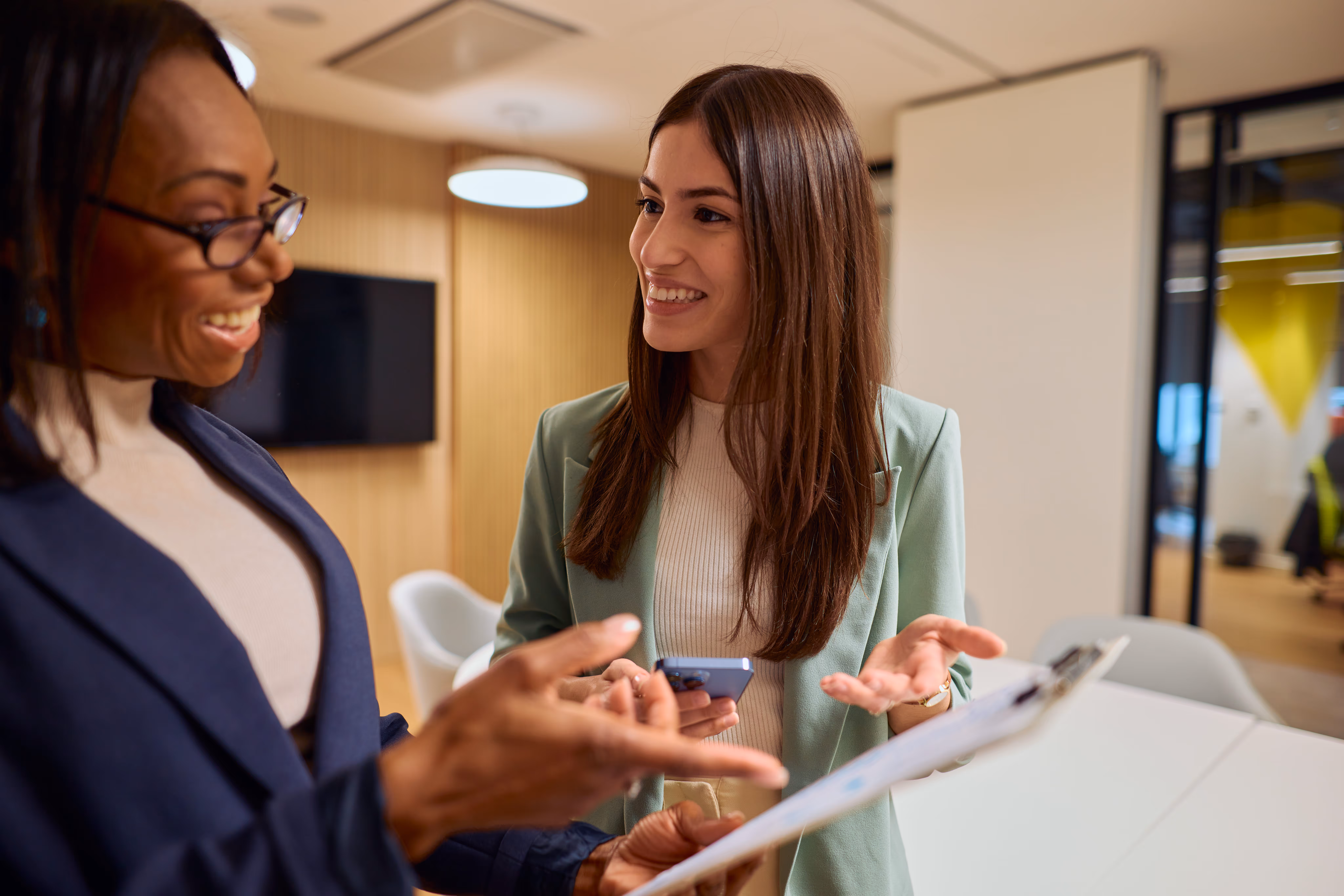 Two professional women smiling and discussing documents on a clipboard in a modern office.