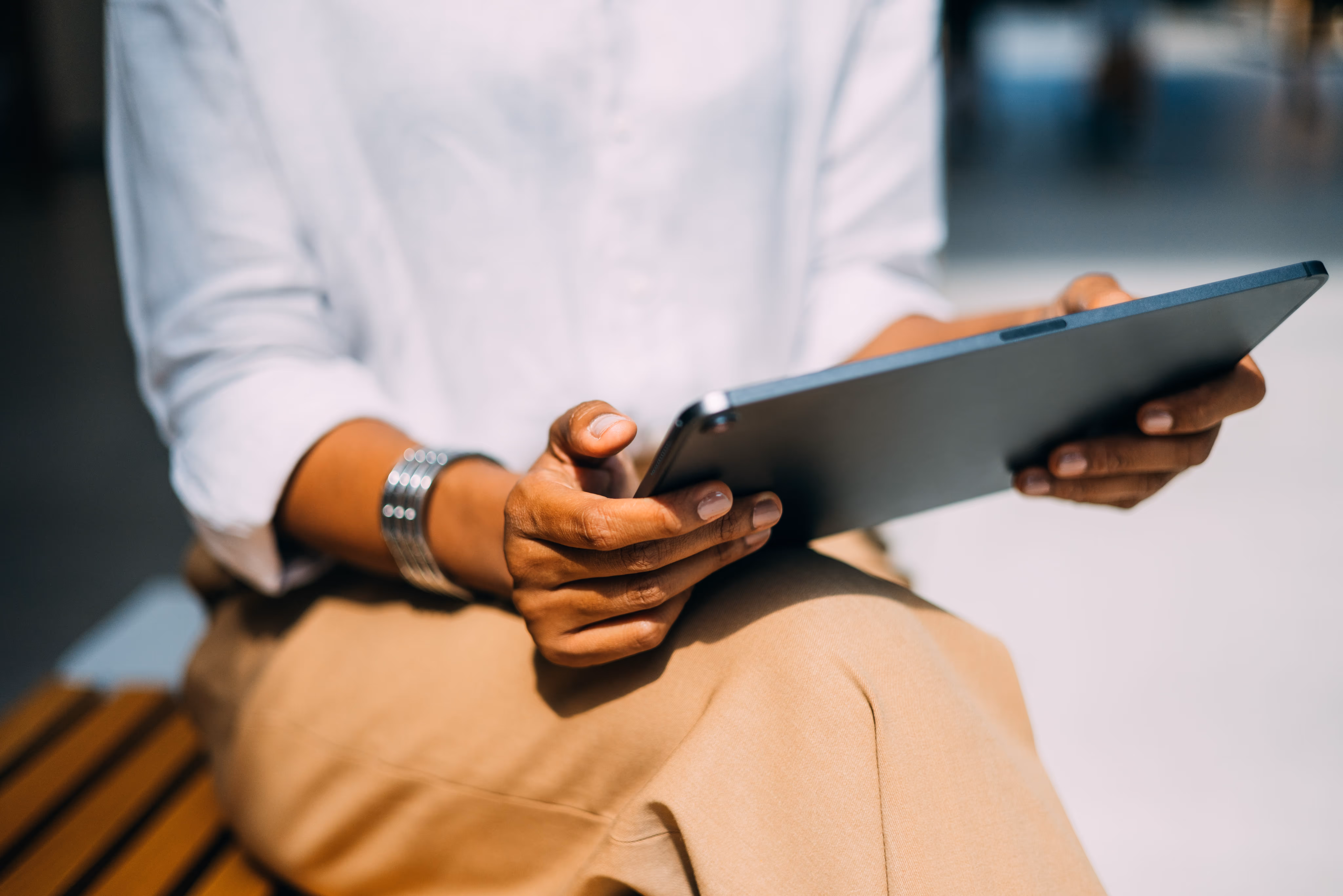 Person wearing a white shirt and beige pants sitting and holding a tablet device.