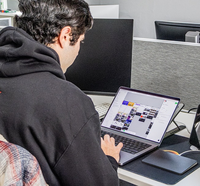 Man in black hoodie working on a laptop displaying a graphic design software at a desk in an office at Tecbot.