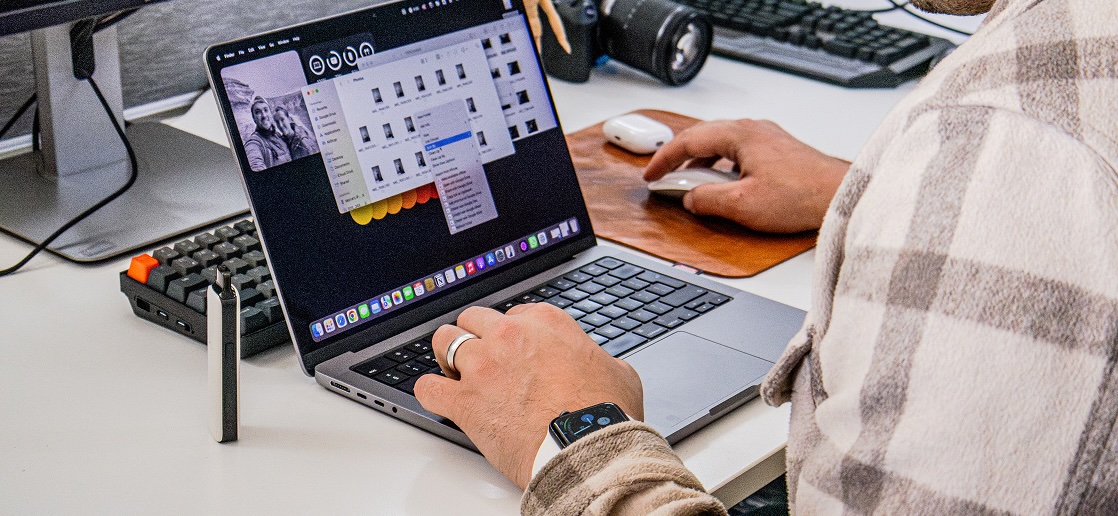 Person using a laptop with photo editing software open, right hand on mouse, wearing a plaid shirt and smartwatch at a white desk with keyboard, and camera at Tecbot.