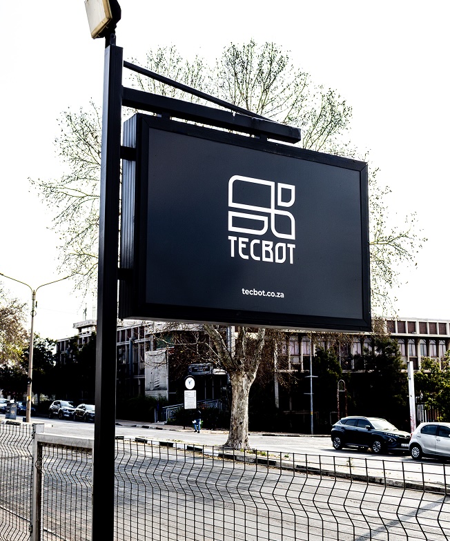 Black signboard mounted on a pole displaying the Tecbot logo and website tecbot.co.za along a street with parked cars and buildings in the background.