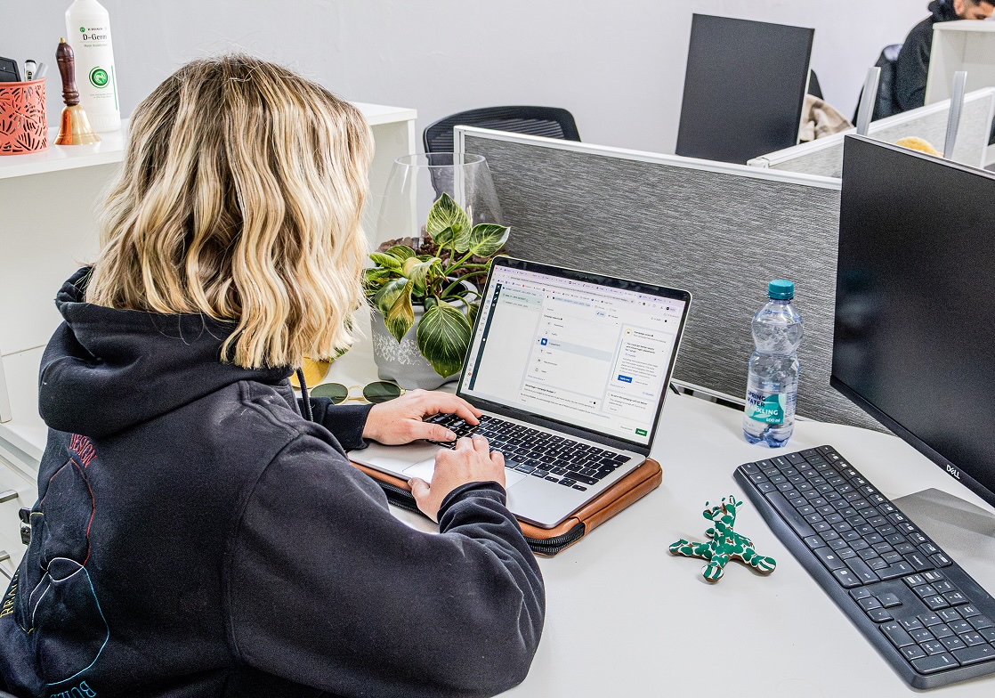 Person with blonde hair in a dark hoodie working on a laptop at a white desk with a plant, water bottle, and keyboard at Tecbot.
