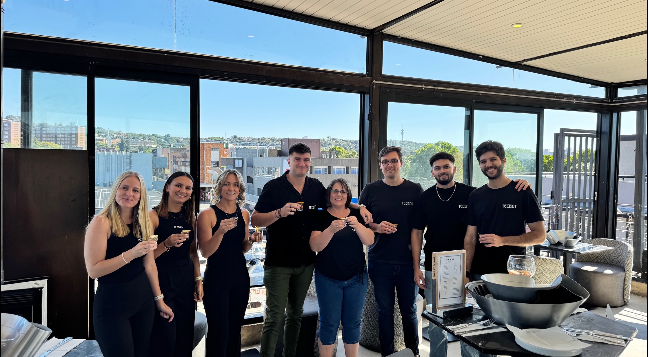 Person with blonde hair in a dark hoodie working on a laptop at a white desk with a plant, water bottle, and keyboard at Tecbot.