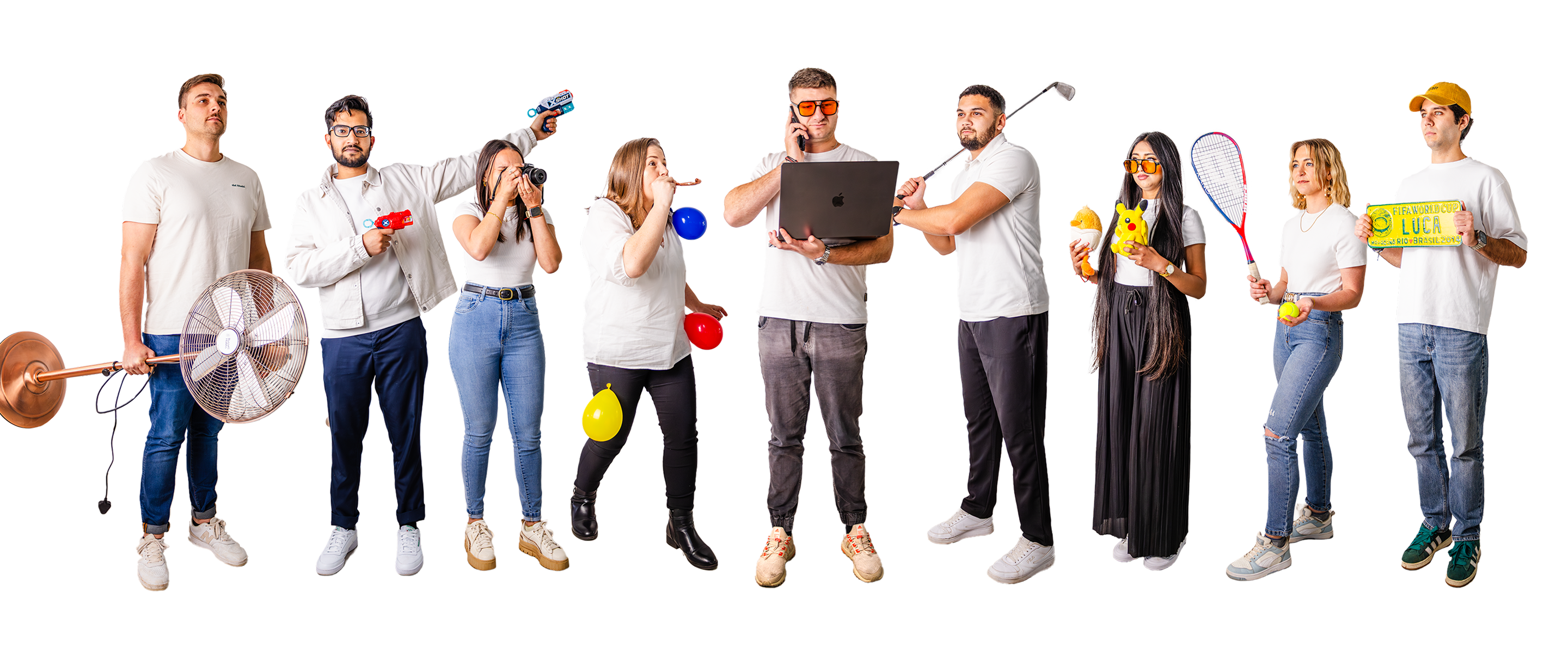 Tecbot team standing in a row, each holding different objects including a large floor fan, camera, balloons, laptop, golf club, plush toys, tennis racket, tennis ball, and a yellow sign, all wearing casual white shirts and jeans or pants.