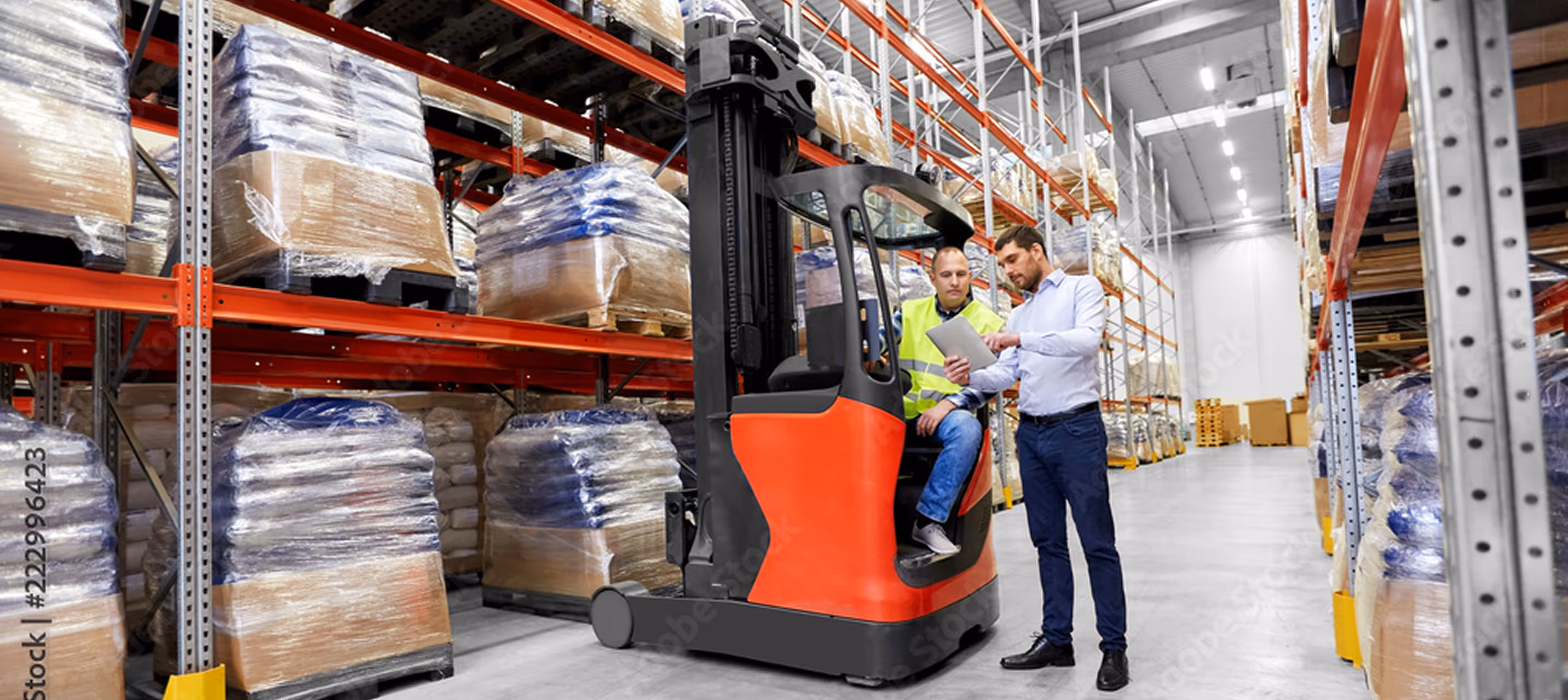 Two workers discuss paperwork in a warehouse, one sitting on a red forklift.