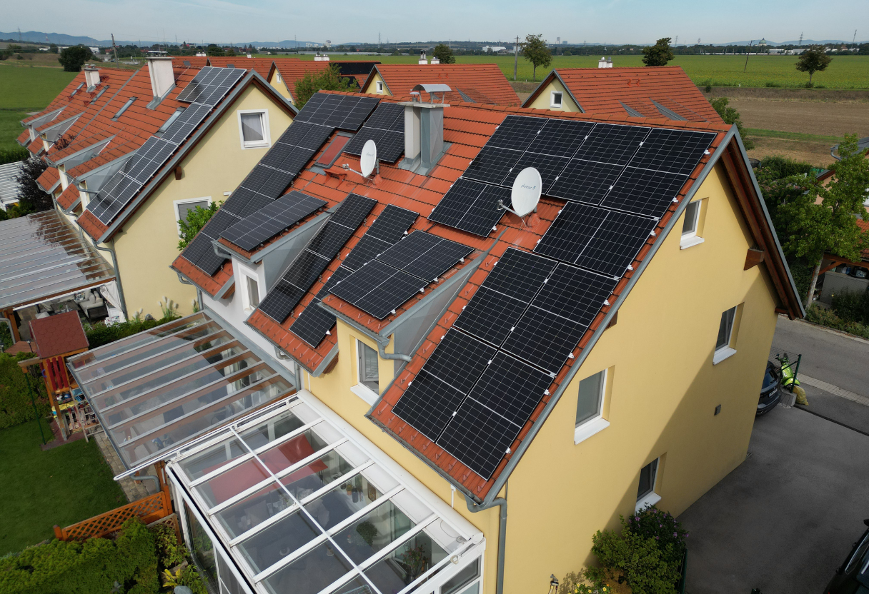 Two yellow houses with red-tiled roofs covered with multiple solar panels, located in a suburban area with green fields in the background.