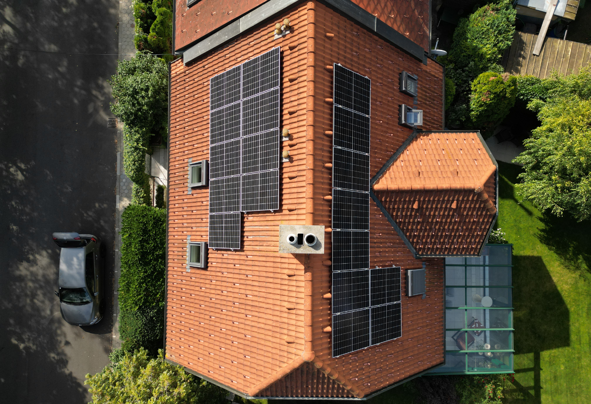 Aerial view of a house roof with solar panels installed and surrounding green garden and street with parked car.
