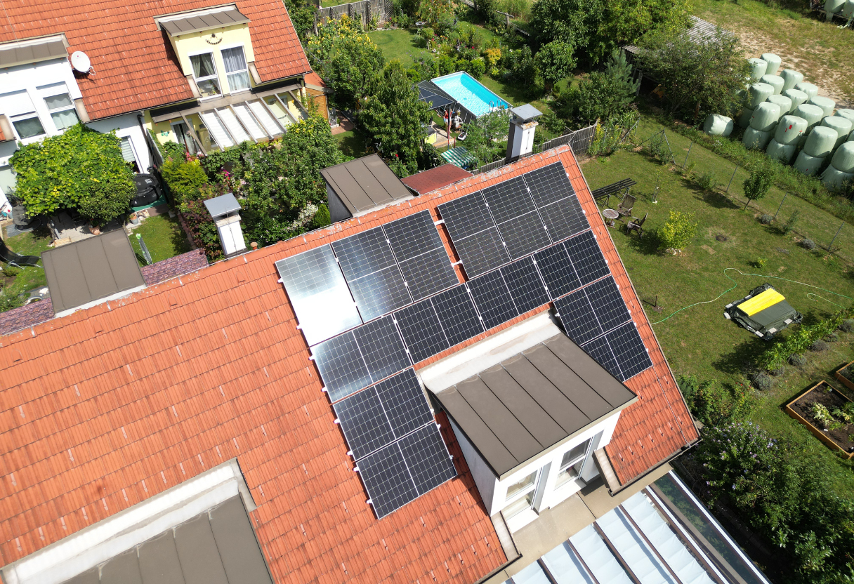 Aerial view of a house with solar panels installed on a red-tiled roof and a green garden with a small swimming pool in the background.