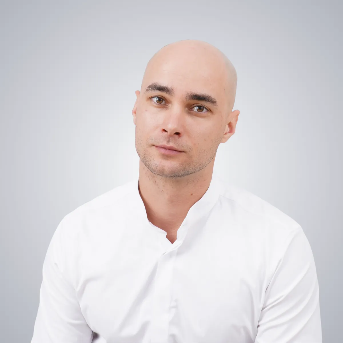 Portrait of a bald man with brown eyes wearing a white collared shirt against a light grey background.