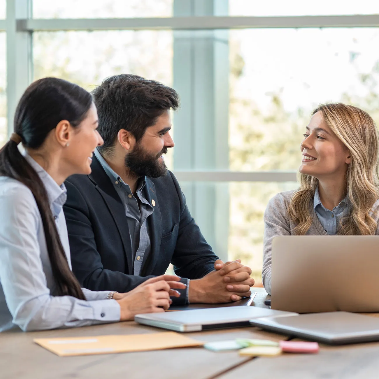 Tre colleghi sorridenti discutono seduti a un tavolo con laptop aperti in un ufficio luminoso.