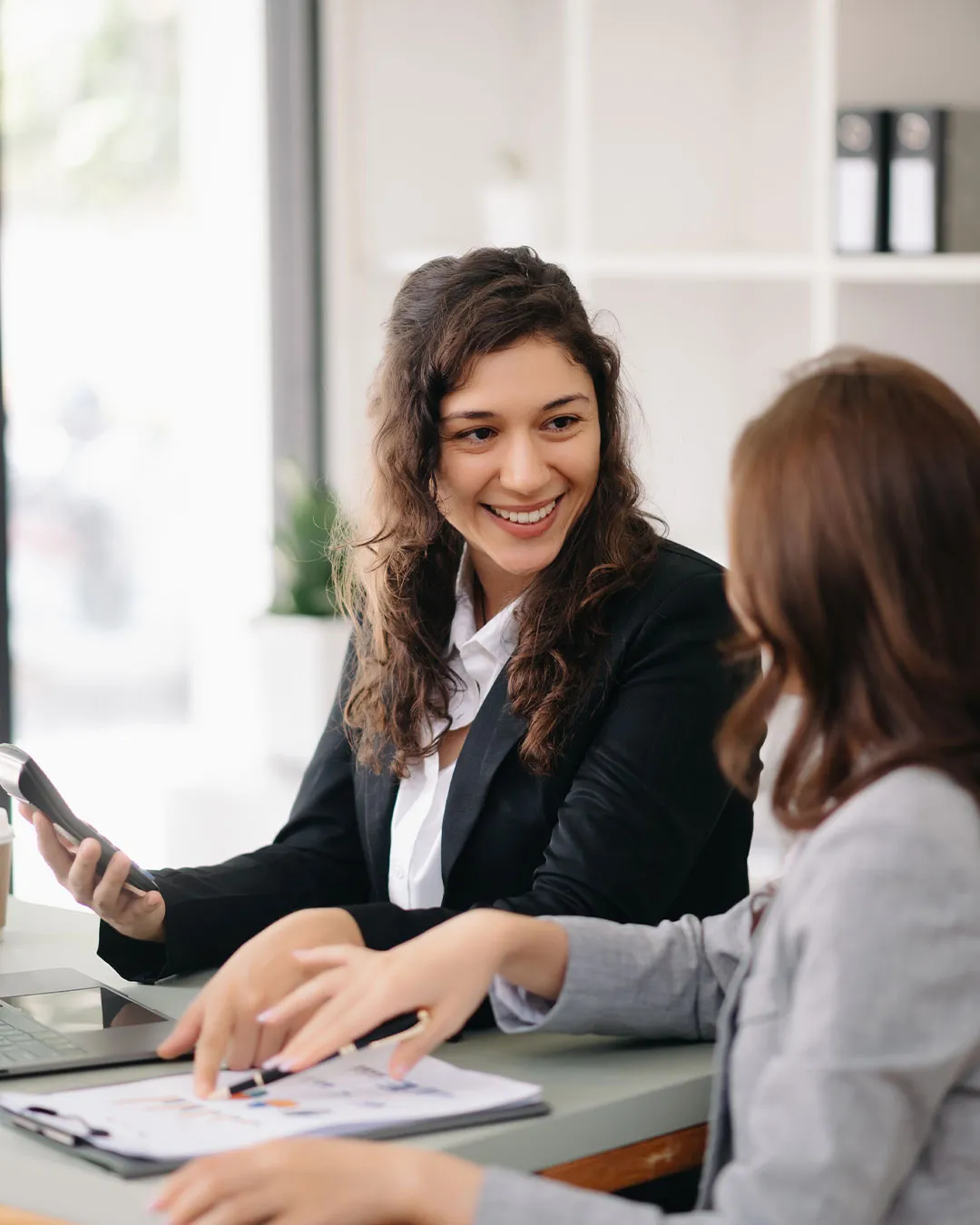 Due donne d'affari sorridenti discutono di un documento con grafici in un ufficio luminoso.