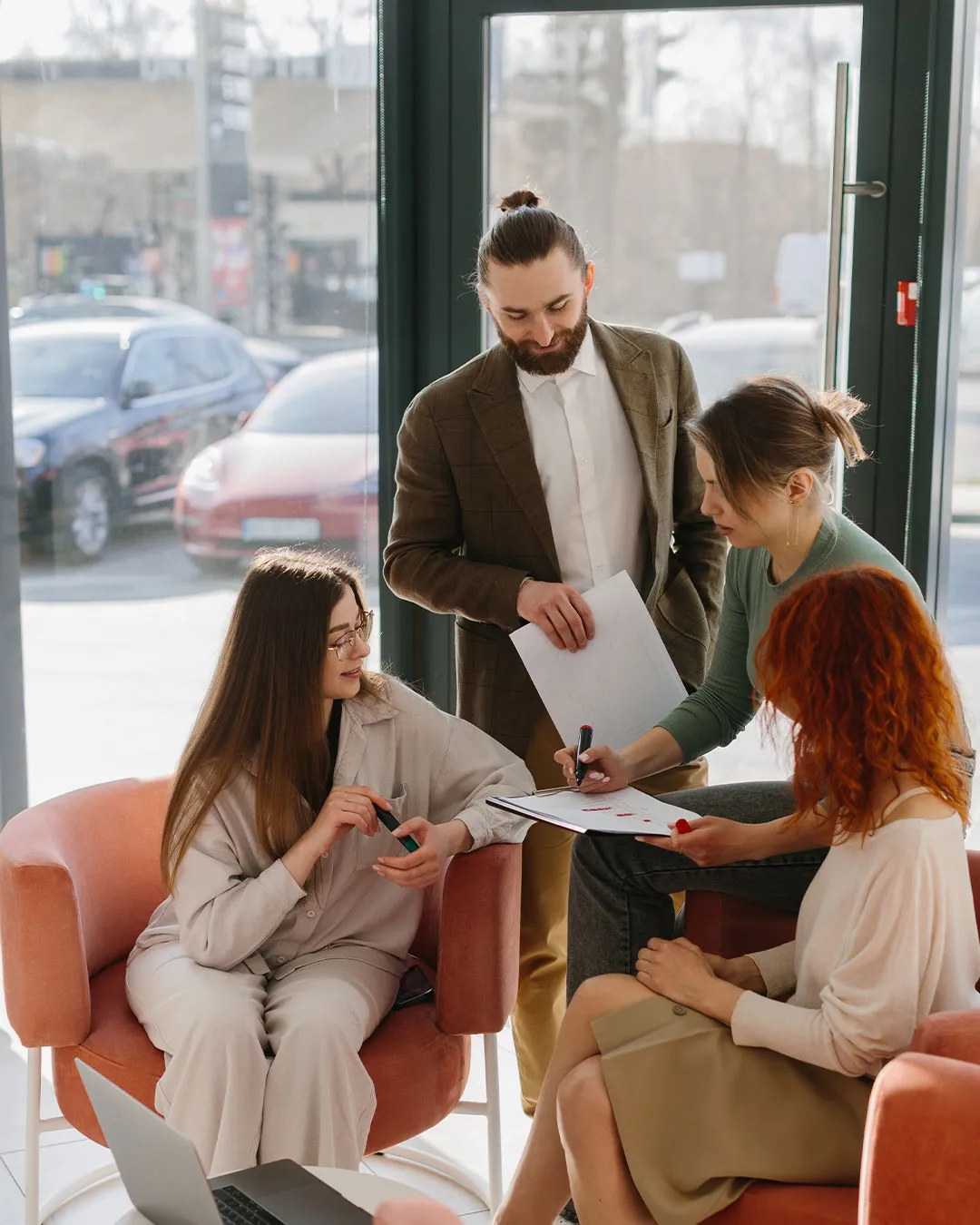 Quattro giovani professionisti che discutono e collaborano seduti su poltrone arancioni vicino a una grande finestra con vista sul parcheggio.