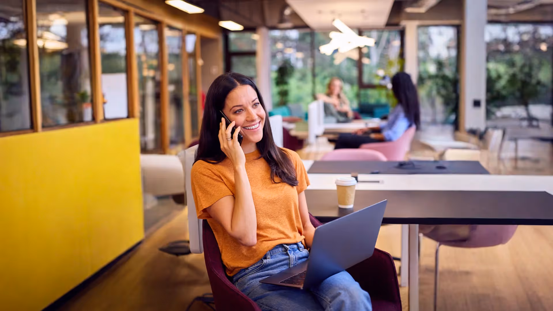 Donna sorridente con maglietta arancione che parla al telefono mentre lavora con un laptop in un ufficio moderno.