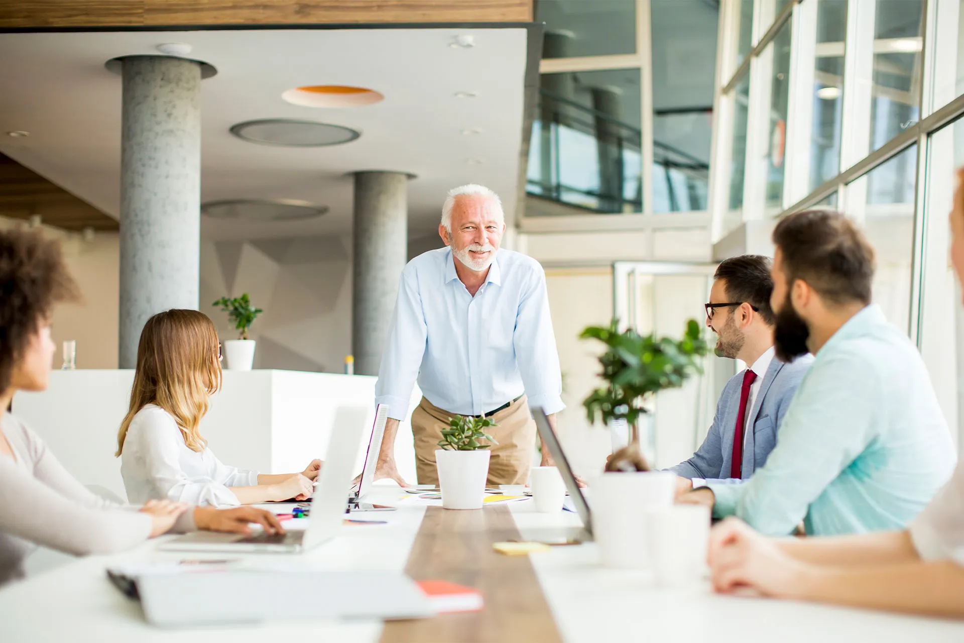 Uomo anziano in camicia azzurra che conduce una riunione in ufficio con colleghi seduti attorno a un tavolo.