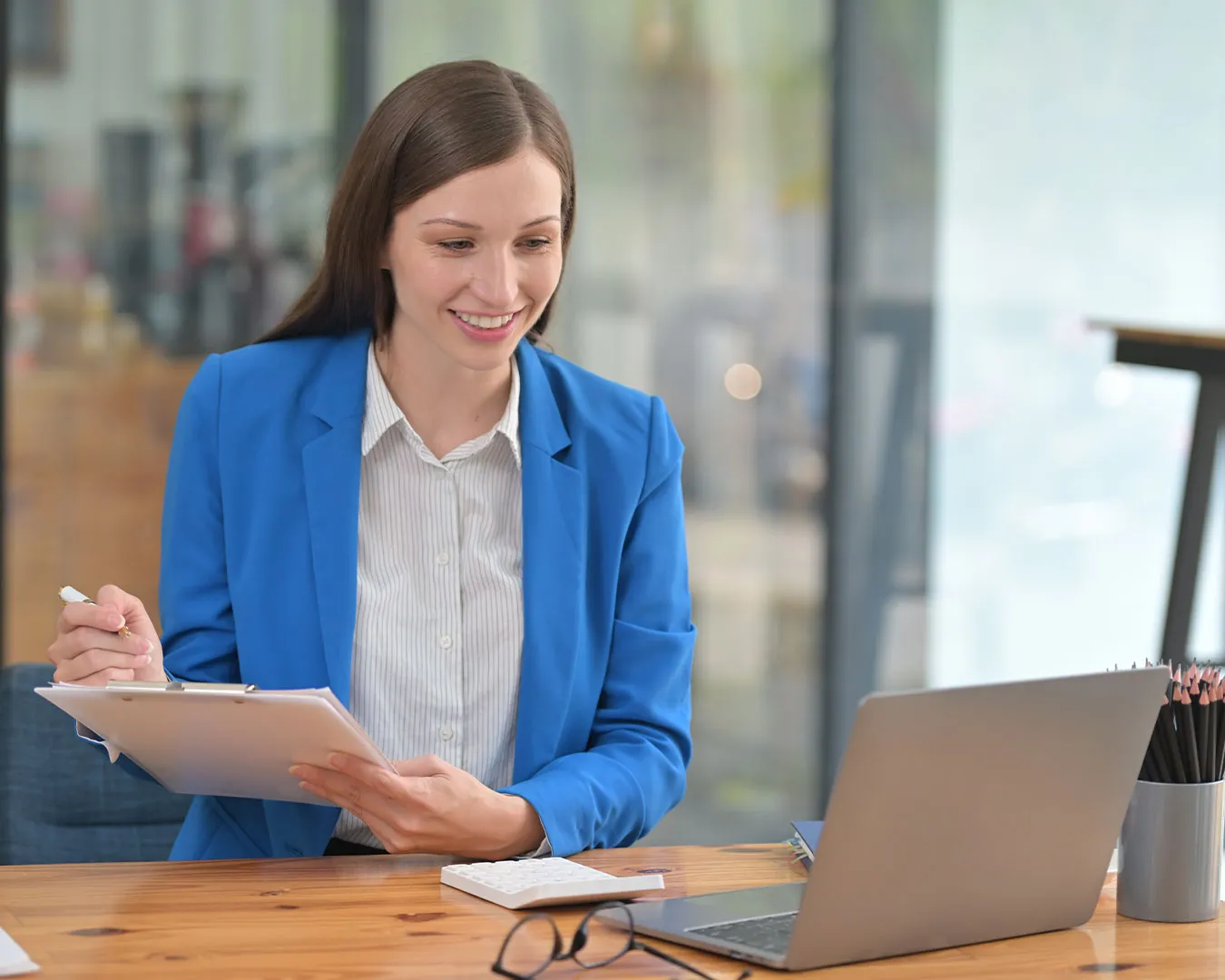 Donna sorridente in giacca blu che tiene una clip e guarda un laptop su una scrivania.