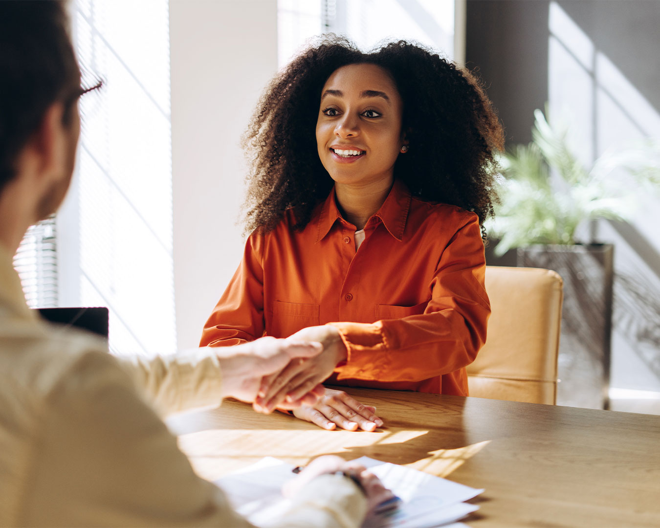 Donna sorridente con camicia arancione che stringe la mano a una persona durante un colloquio di lavoro in un ufficio luminoso.