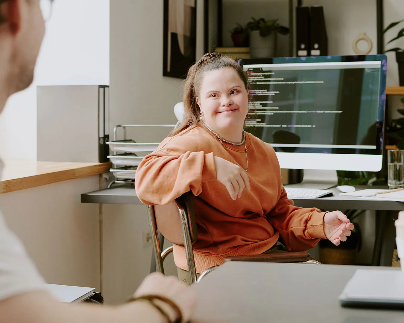Donna con sindrome di Down seduta a una scrivania in ufficio con un computer desktop che mostra codice, mentre conversa con una persona non visibile in primo piano.
