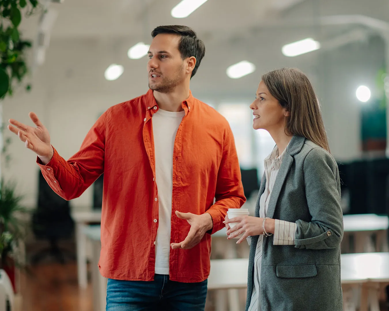 Due colleghi discutono in ufficio, un uomo con camicia arancione parla mentre una donna con giacca grigia tiene una tazza di caffè.