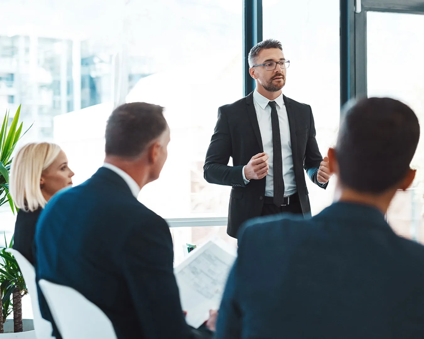 Uomo in abito scuro che parla in piedi davanti a un gruppo di persone in un ufficio luminoso.