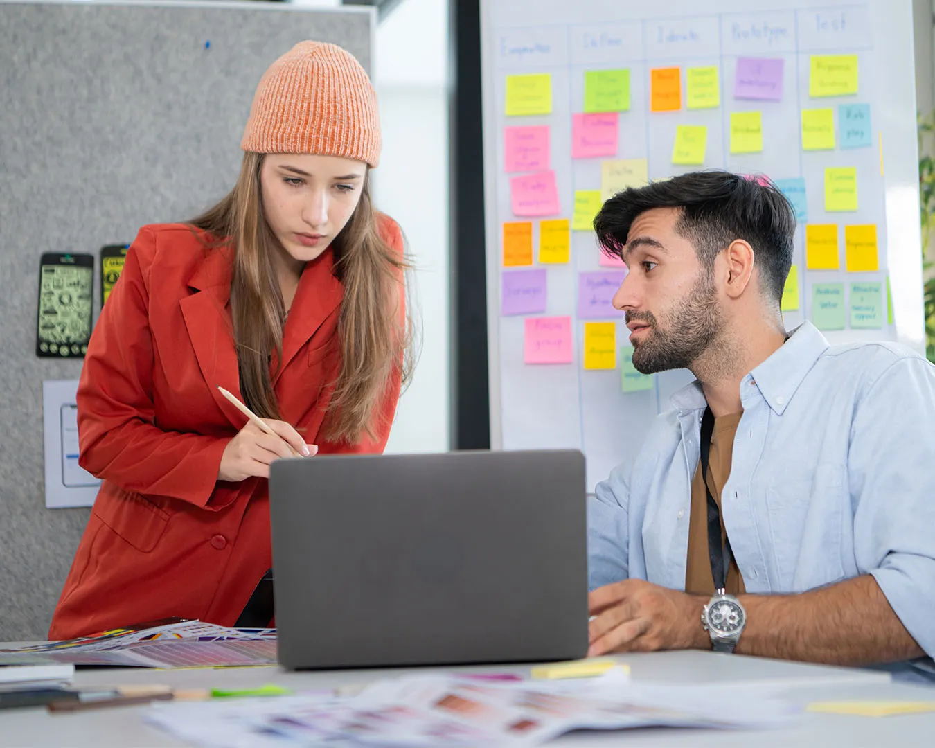 Due colleghi discutono di lavoro davanti a un laptop con una lavagna di post-it colorati alle spalle.
