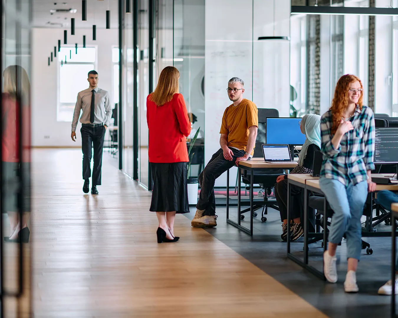 Persone in ufficio moderno con pavimento in legno, alcuni seduti e altri in piedi che conversano e lavorano ai computer.