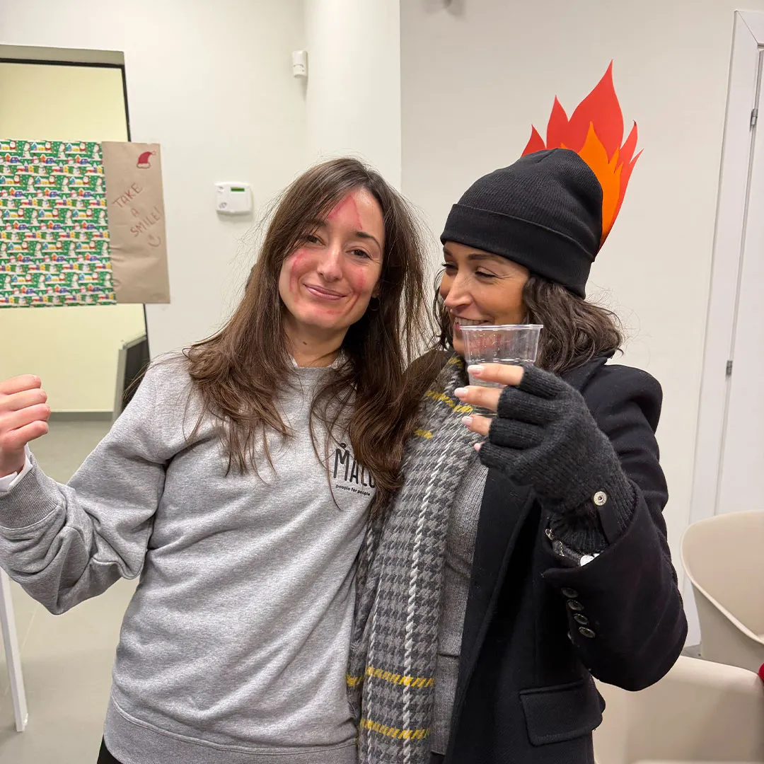 Due donne sorridenti abbracciate, una con maglione grigio e segni di trucco rosso sul viso, l'altra con berretto nero decorato con fiamme di carta e guanti neri, che tiene un bicchiere di plastica.