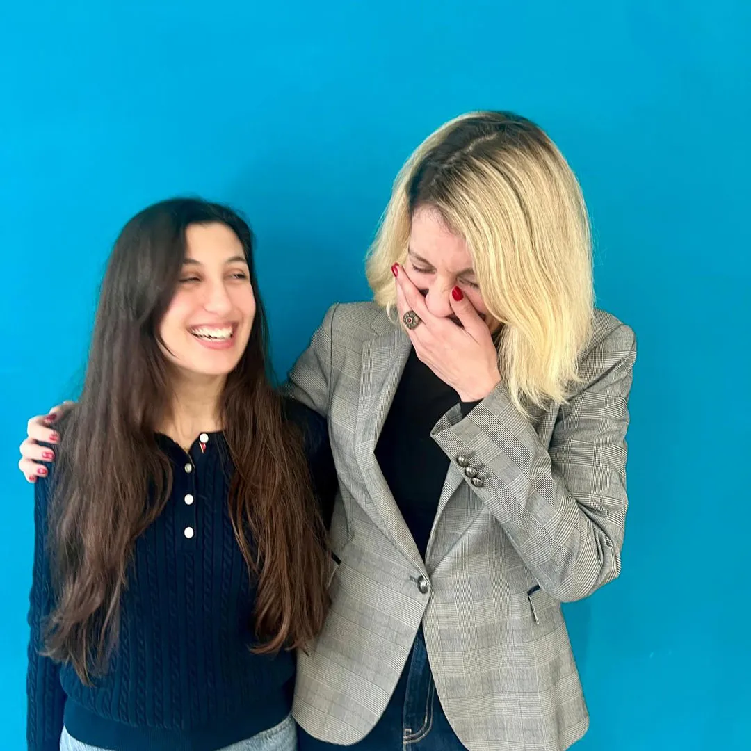 Due donne sorridenti, una con capelli lunghi castani e maglione blu, e l'altra con capelli biondi che ride coprendosi la bocca, davanti a uno sfondo blu.