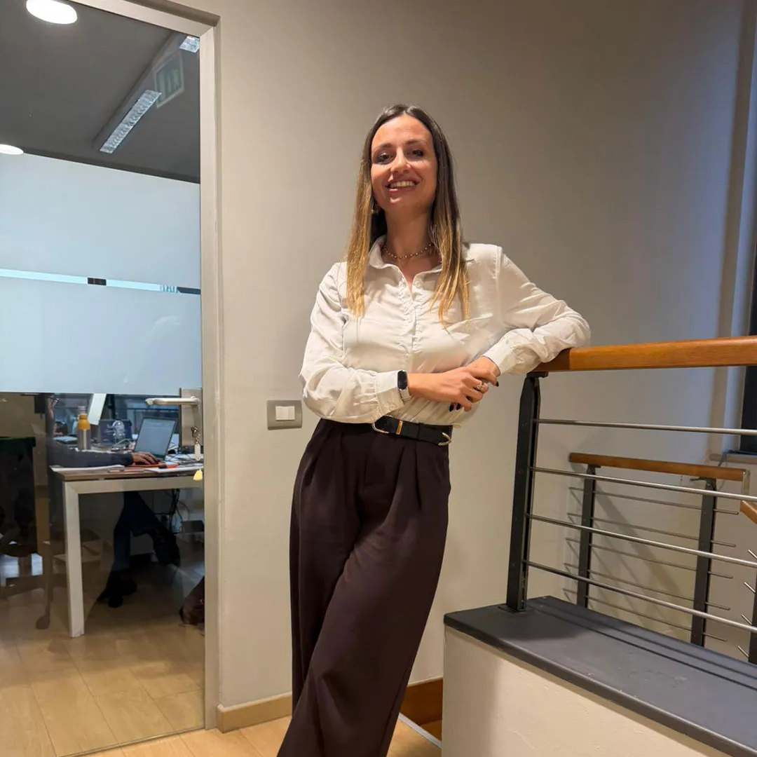 Donna sorridente con camicia bianca e pantaloni neri che si appoggia a una ringhiera in un ufficio moderno.