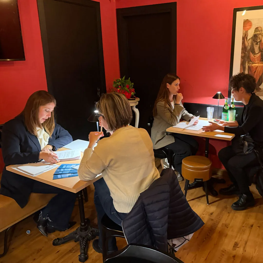 Quattro donne sedute a due tavoli in un ambiente con pareti rosse, impegnate in discussioni e letture di documenti.