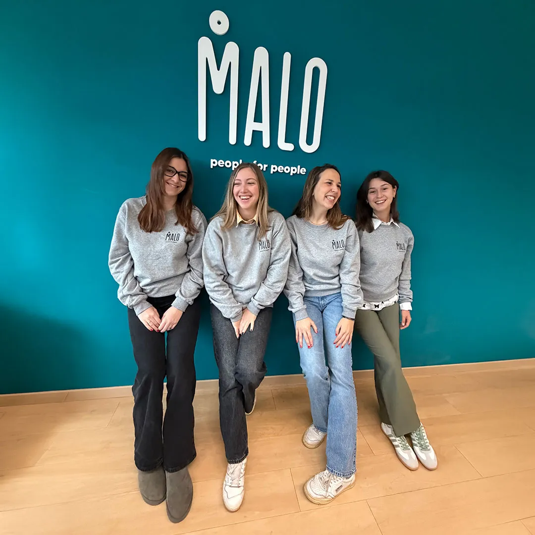 Quattro donne sorridenti sedute davanti a una parete turchese con il logo bianco di MALO e la scritta 'people for people'.