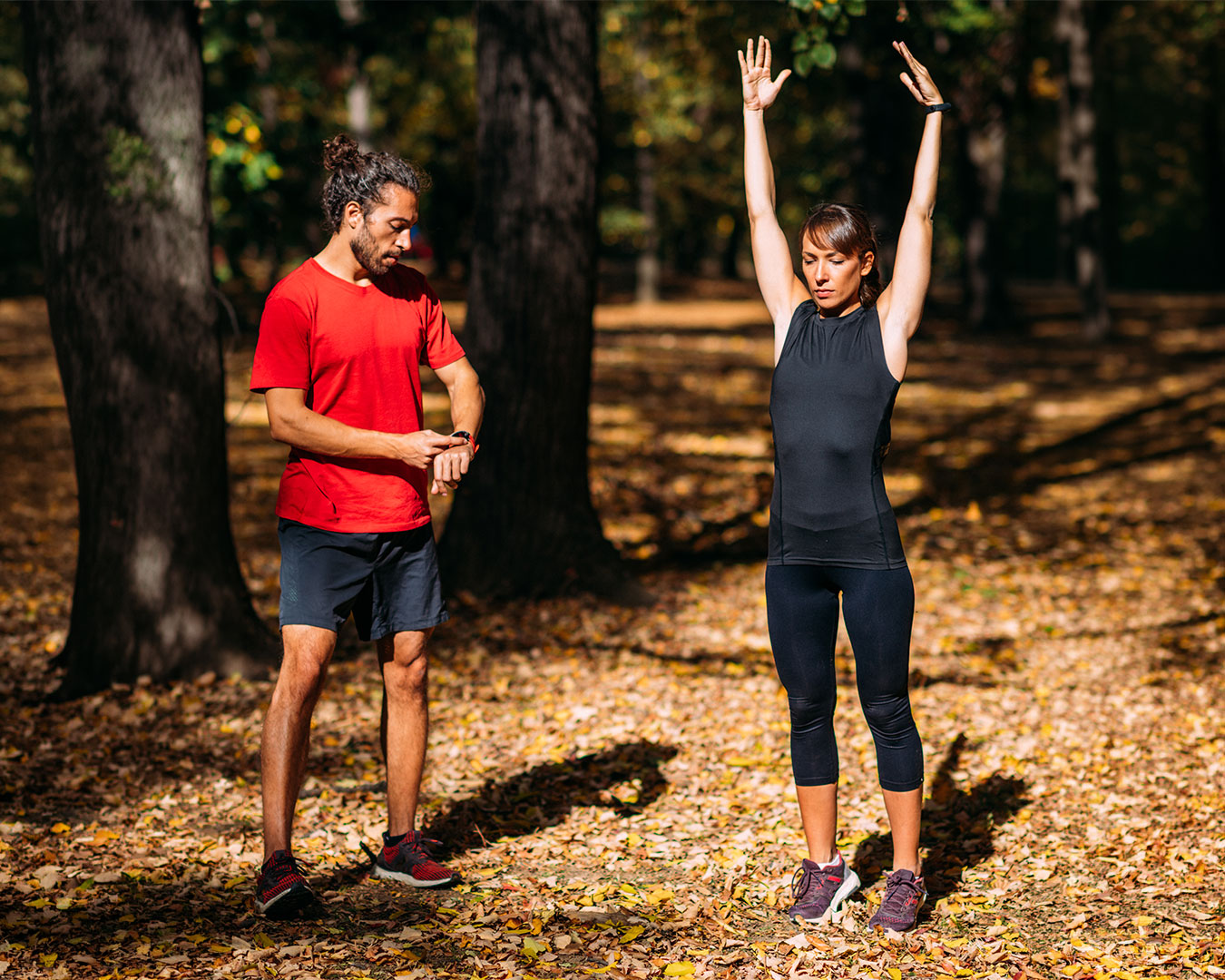 Donna in abbigliamento sportivo con le braccia alzate e uomo che controlla lo smartwatch durante l’allenamento all’aperto in un parco autunnale.